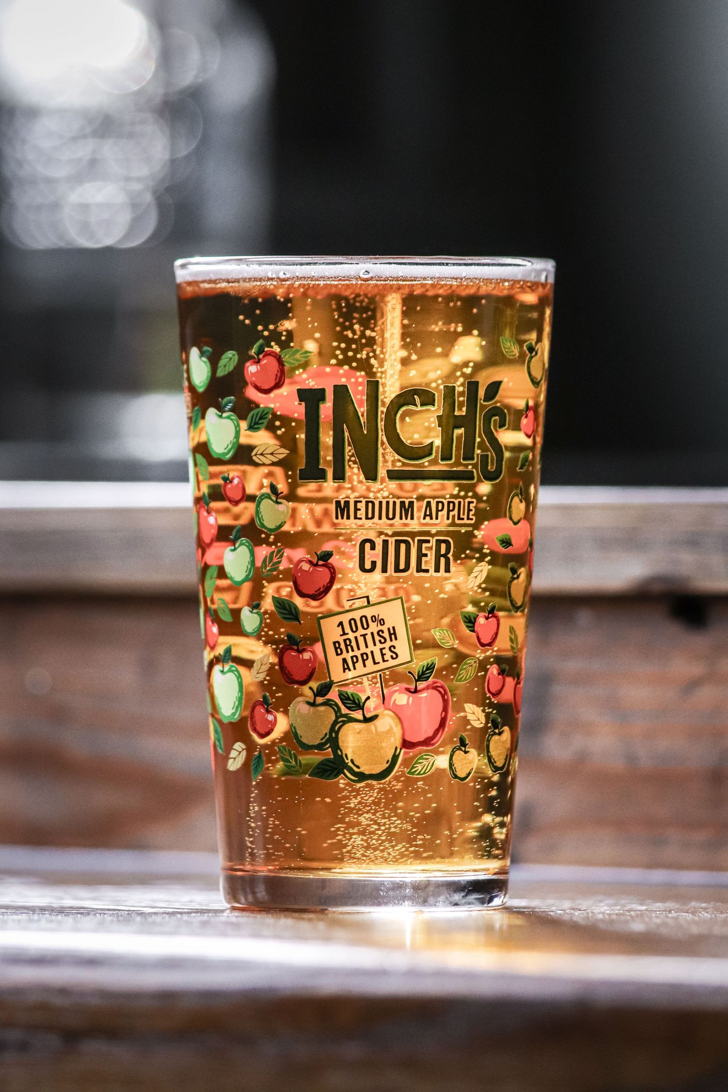 A glass of Inch's medium dry cider with decorative apple illustrations on the glass sits on a wooden surface.