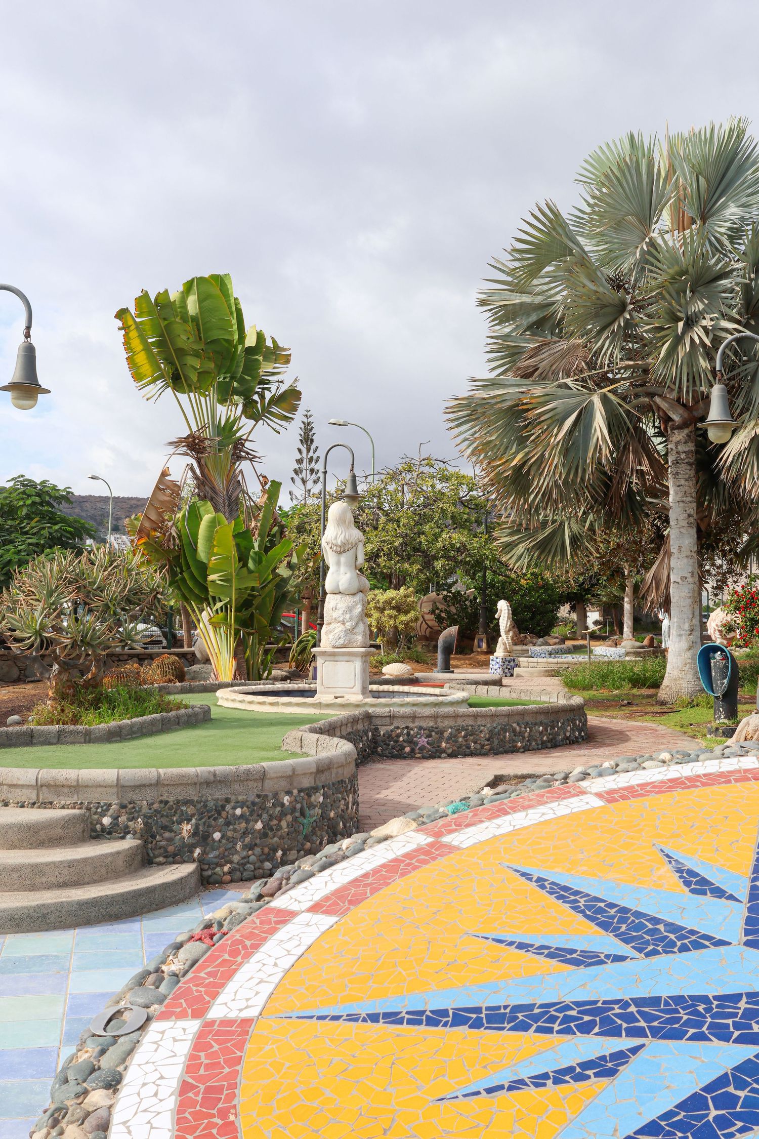 Colorful mosaic tile pattern with geometric designs decorates an outdoor garden pathway surrounded by palm trees and plants.