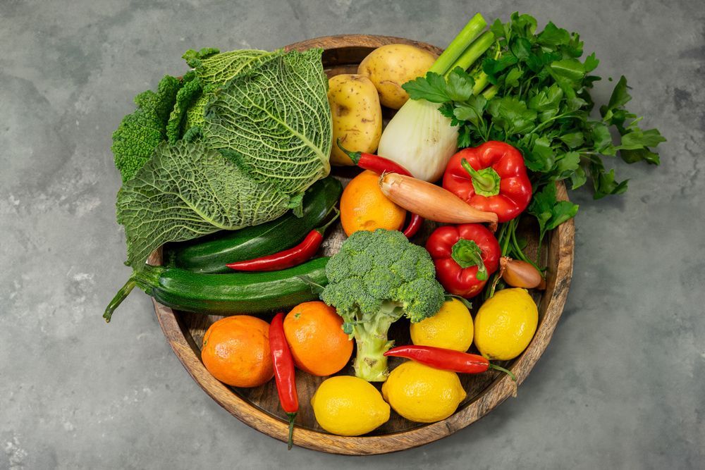 Fresh colorful produce including citrus fruits, vegetables, and leafy greens arranged in wooden bowls on a gray surface.