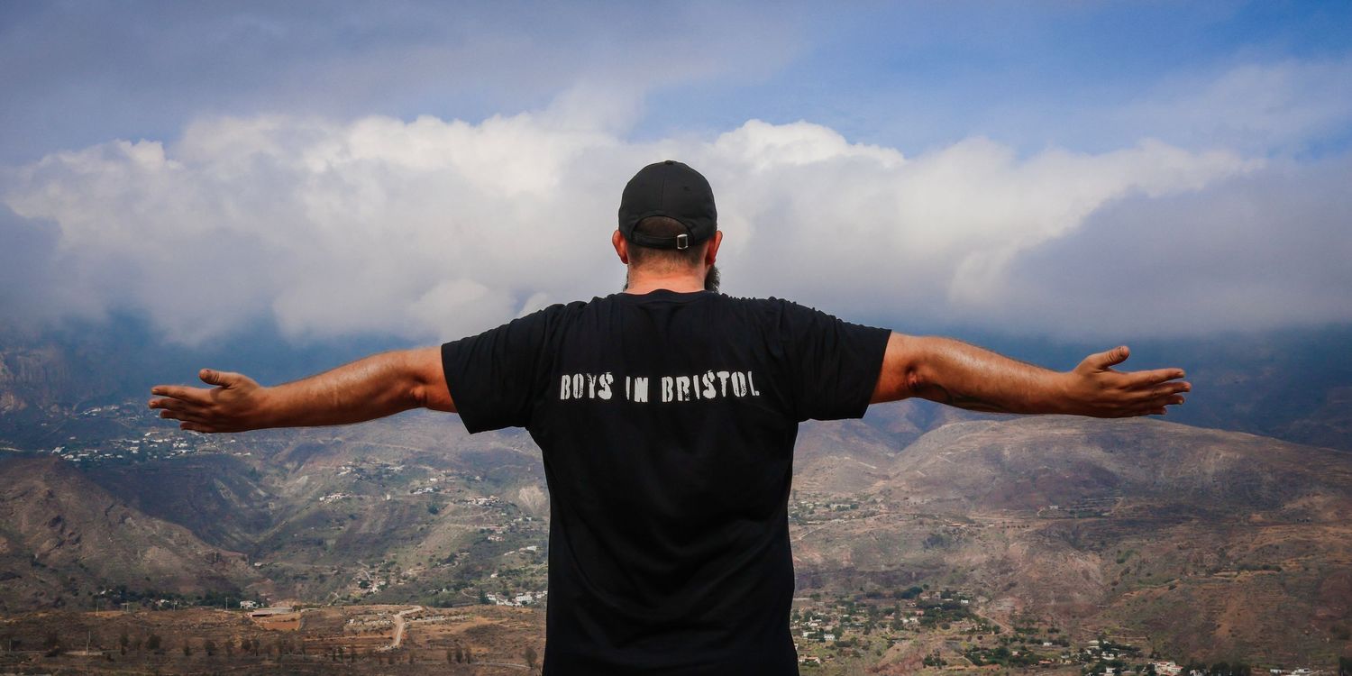 A person stands with arms outstretched overlooking a scenic mountain vista on a cloudy day.
