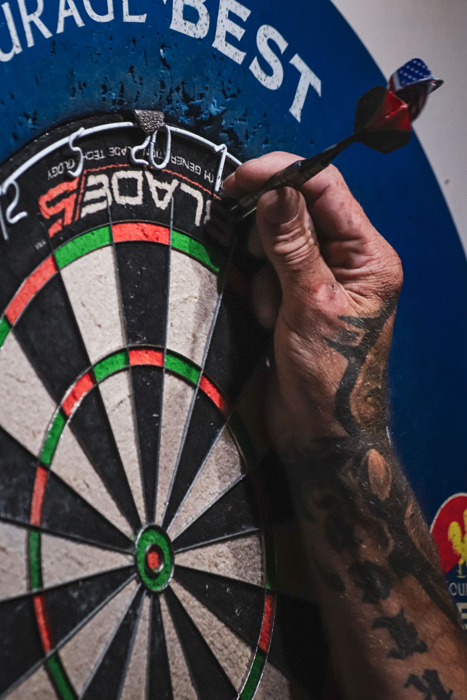A tattooed arm removes a dart from a professional dartboard with red, green and white sections.