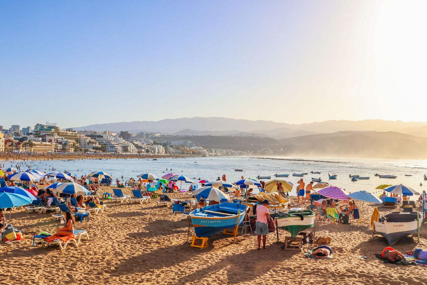 Colorful beach umbrellas and crowds dot the sandy shoreline at sunset along the California coast.