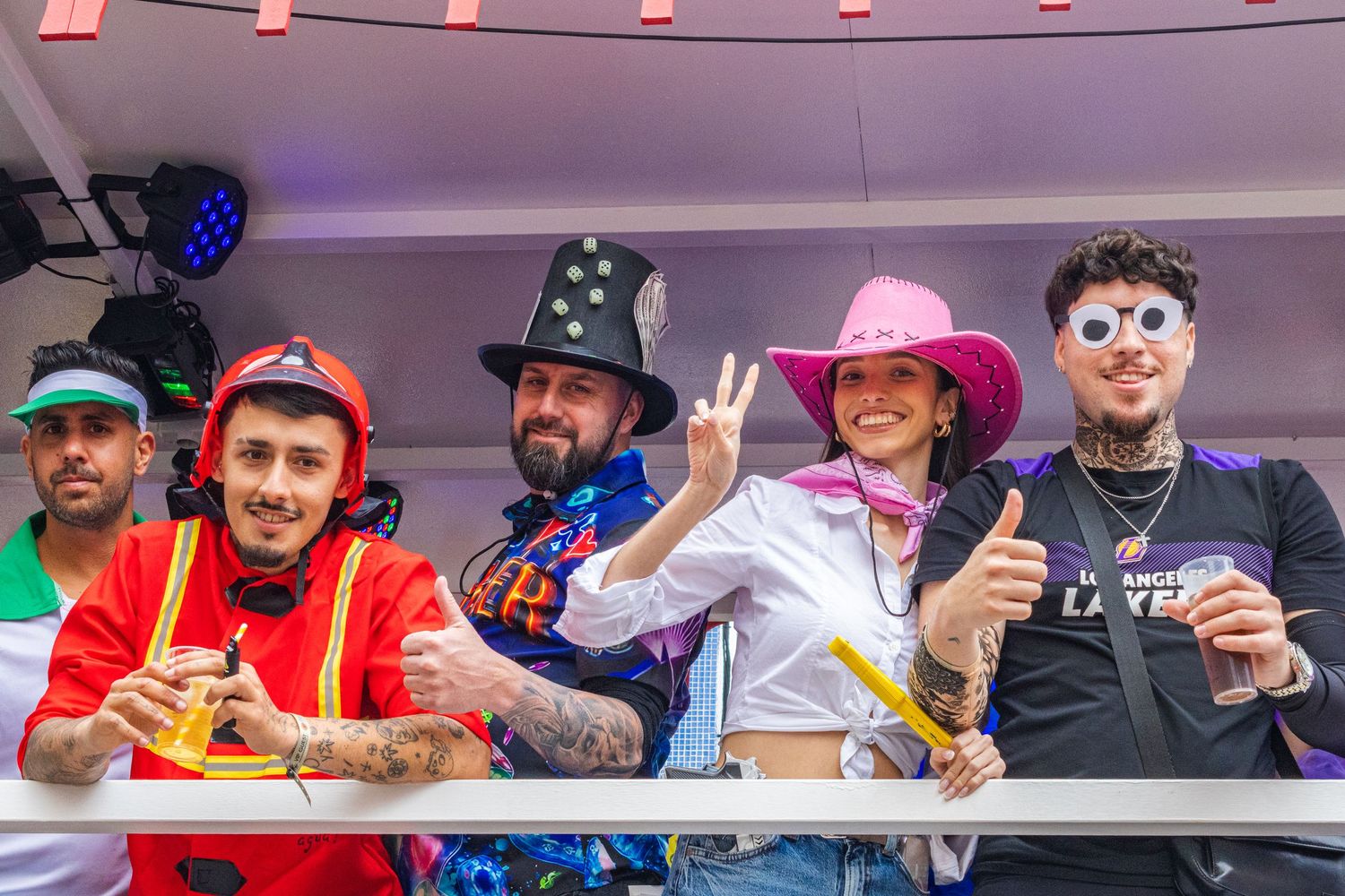 A group of people in colorful hats and costumes pose together at an event showing peace signs and smiling.