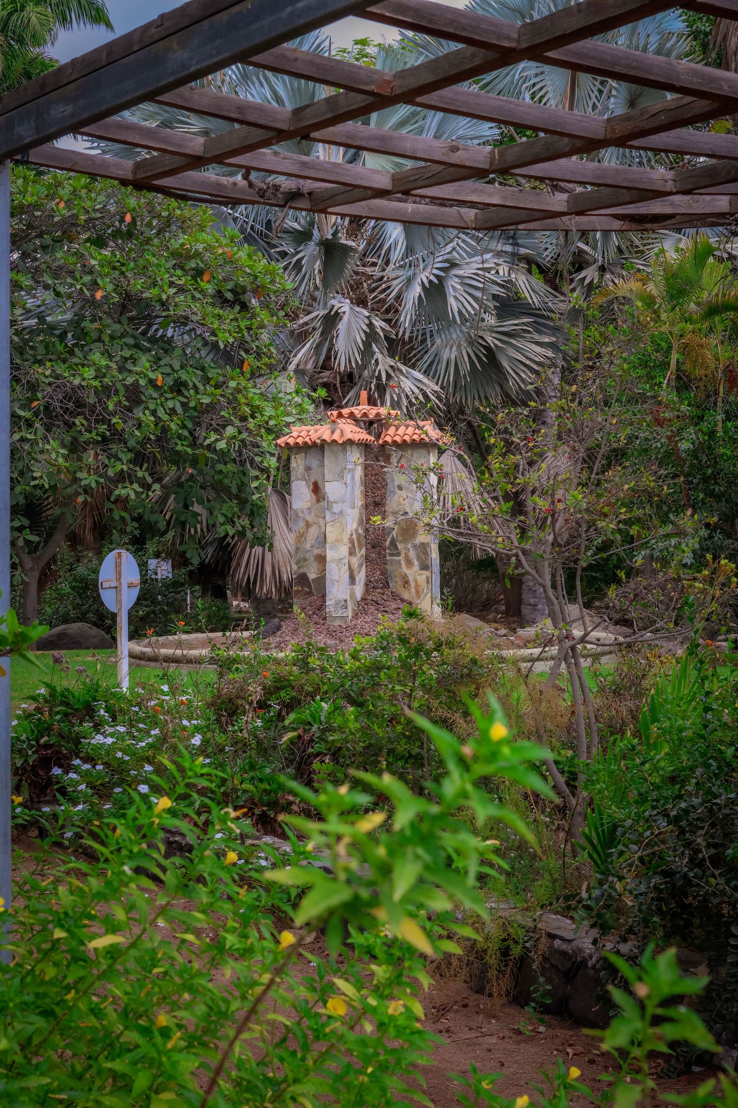 Lush tropical garden pathway with palm trees and rustic pergola leading to a Mediterranean-style stone structure.