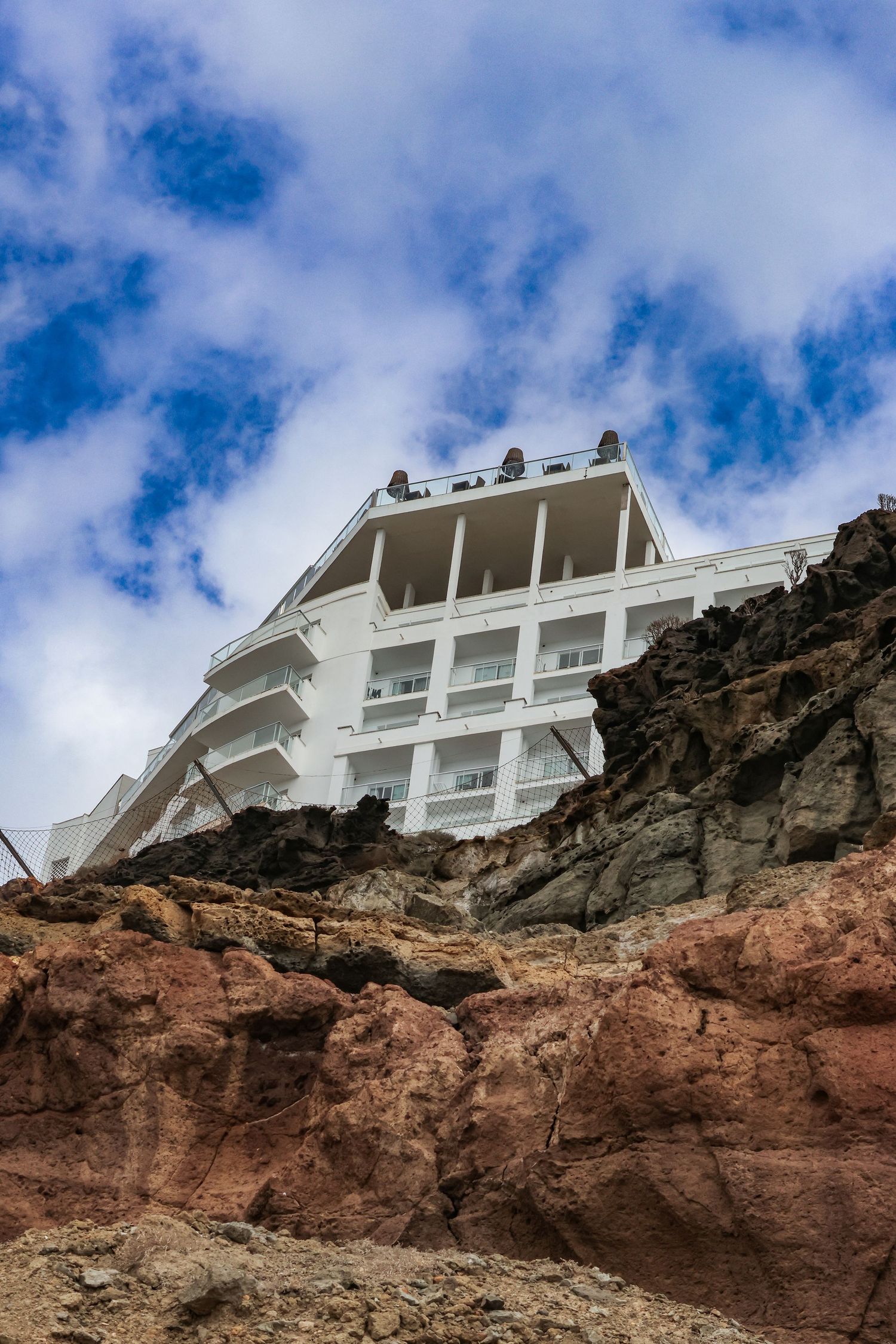 White modern house perched precariously on the edge of red rocky cliffs against a blue cloudy sky.
