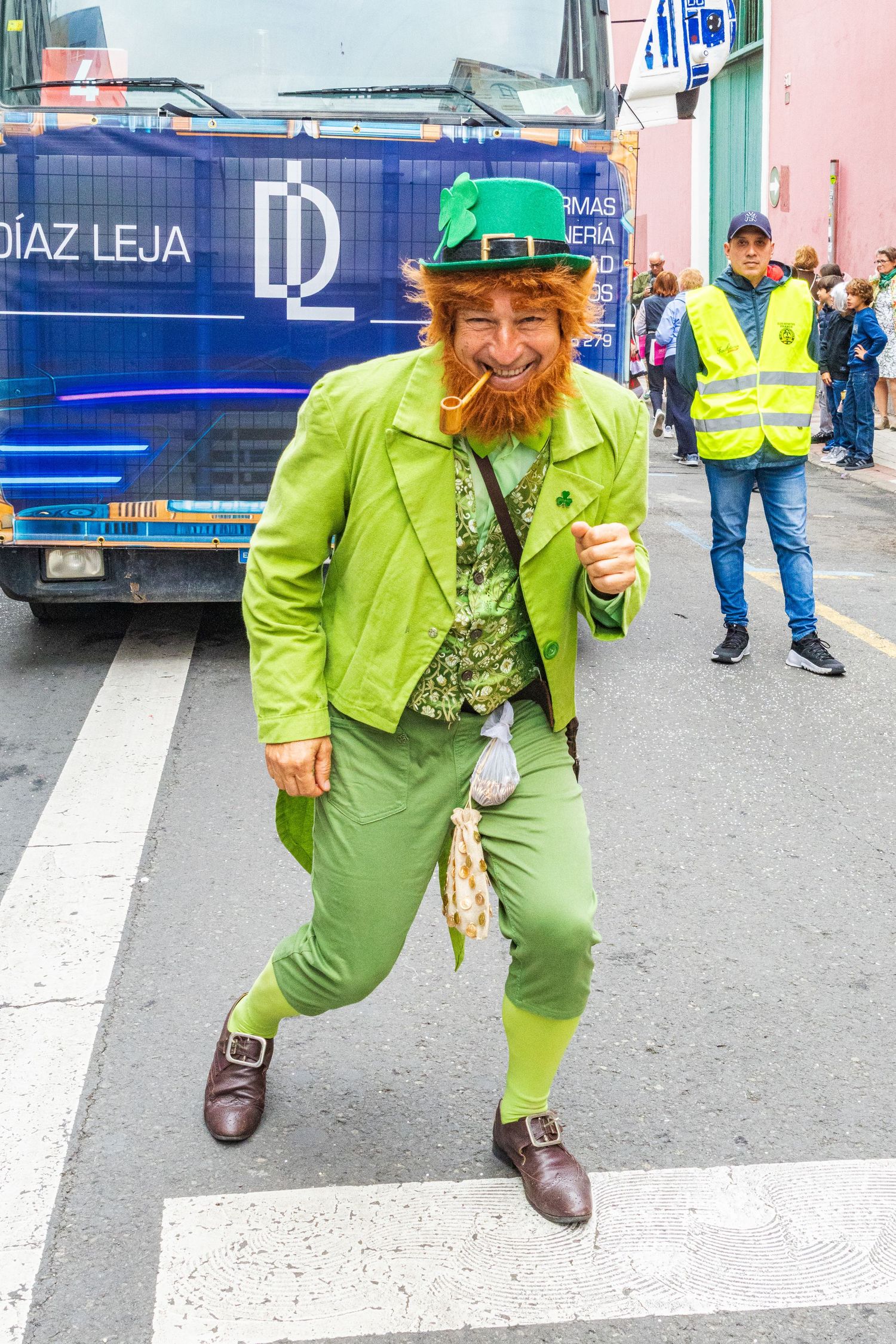 A person in bright green attire and hat performs playful poses on a street near delivery vehicles and workers.