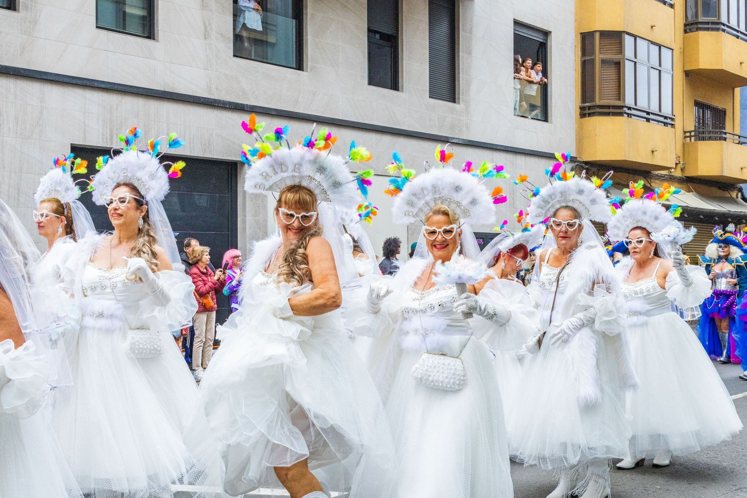 A large group of people dressed in white outfits dance and celebrate during a festive parade or street celebration.