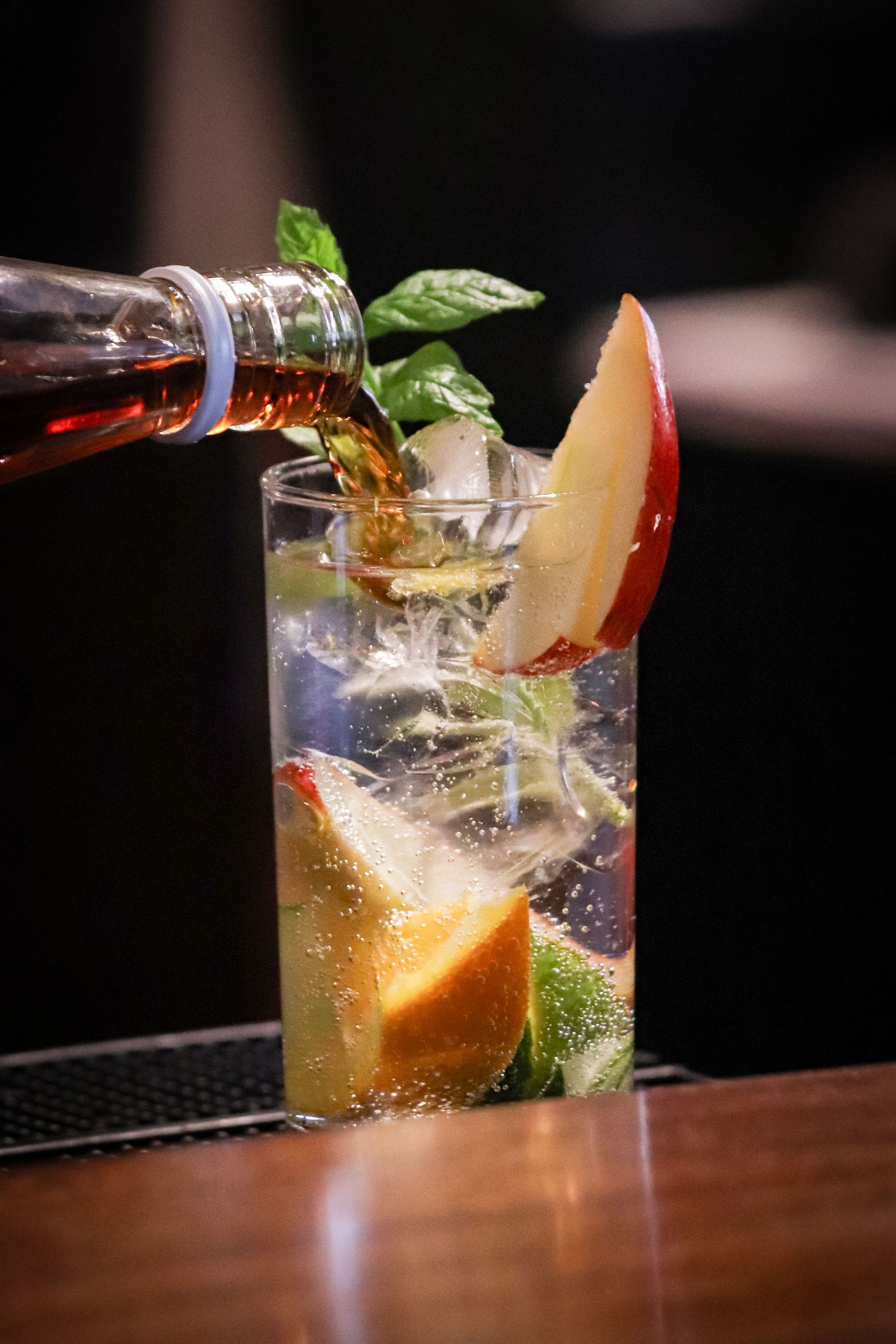 Close-up sequence of pouring a refreshing citrus cocktail with fresh herbs and ice in a glass at a dark bar.