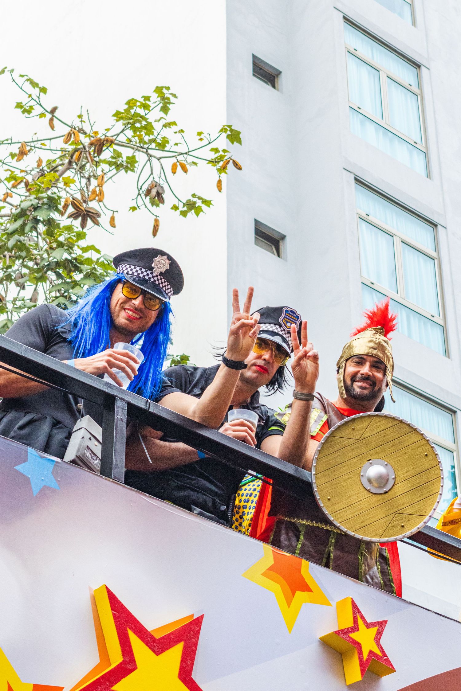 Costumed revelers in colorful outfits wave and celebrate from a balcony decorated with red and yellow stars.