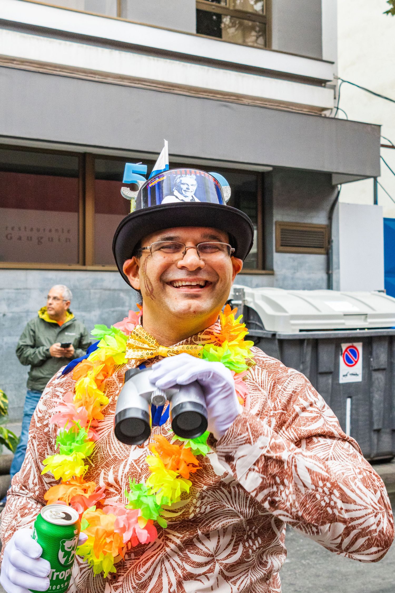 A festive carnival goer wearing a colorful lei and hat with drink holders celebrates at a street party.