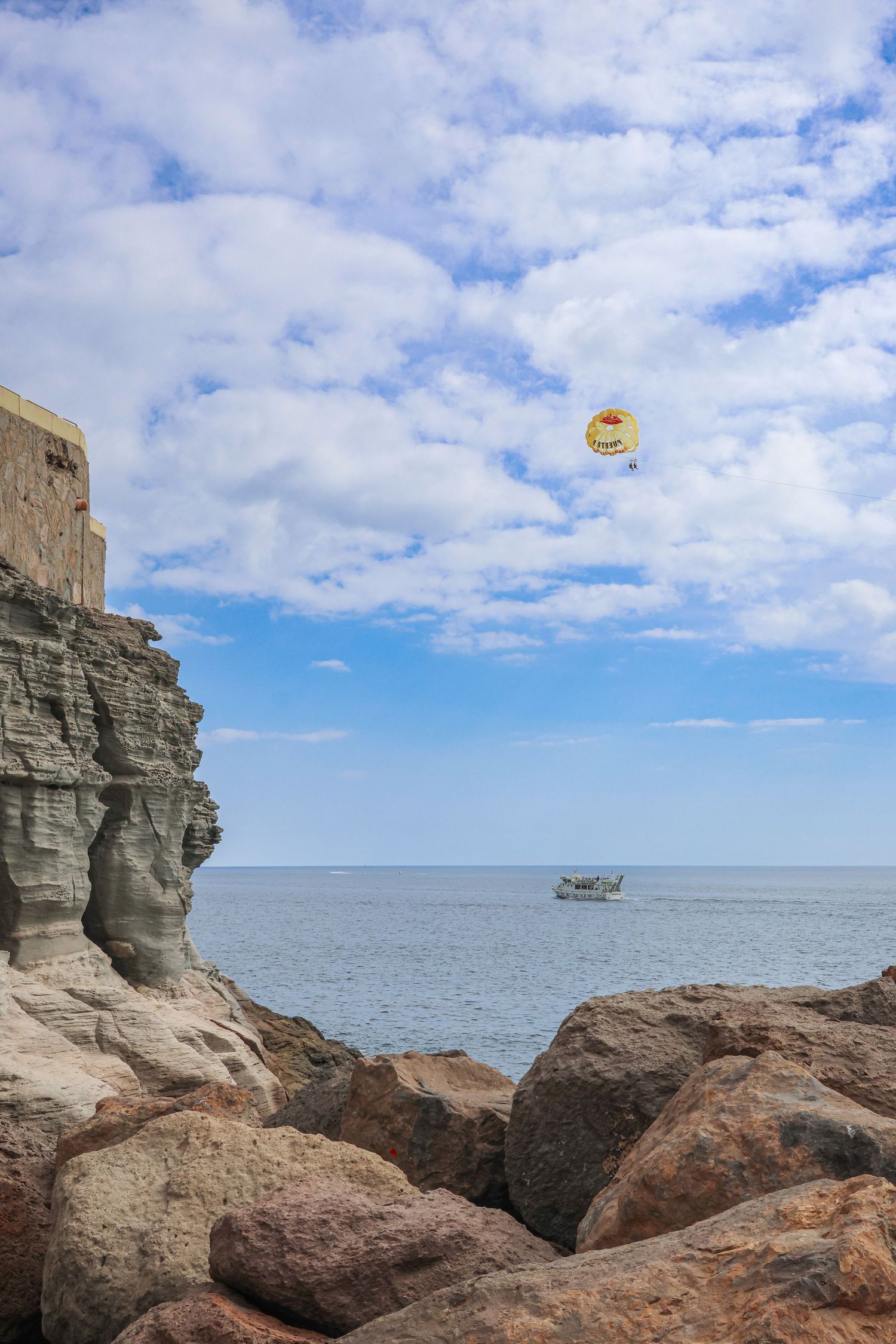 Parasailing activity over ocean waters near rocky coastal cliffs with dramatic cloudy sky backdrop.