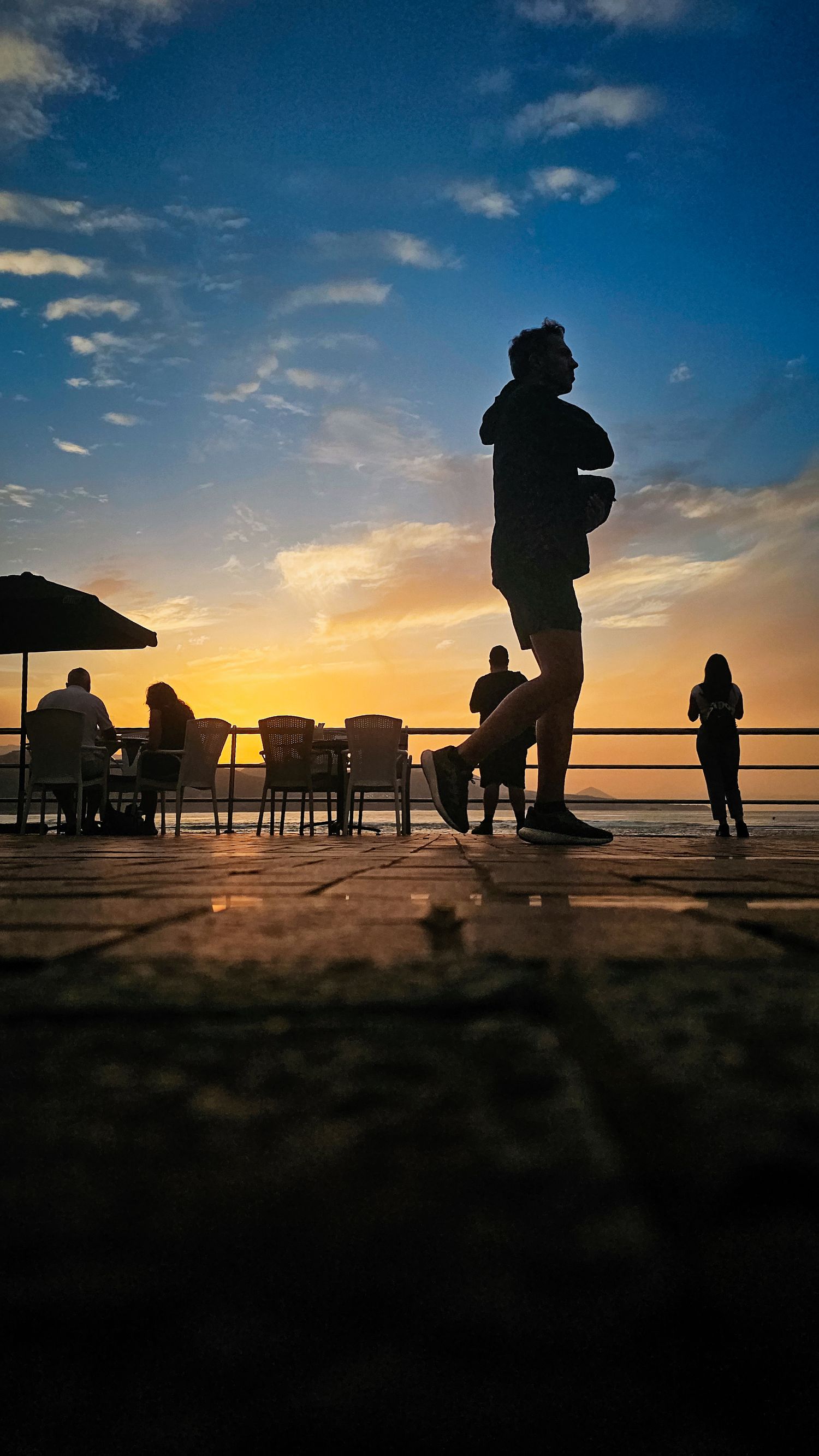 Silhouettes of people enjoying sunset on a pier with dramatic orange sky and clouds reflected on calm waters.