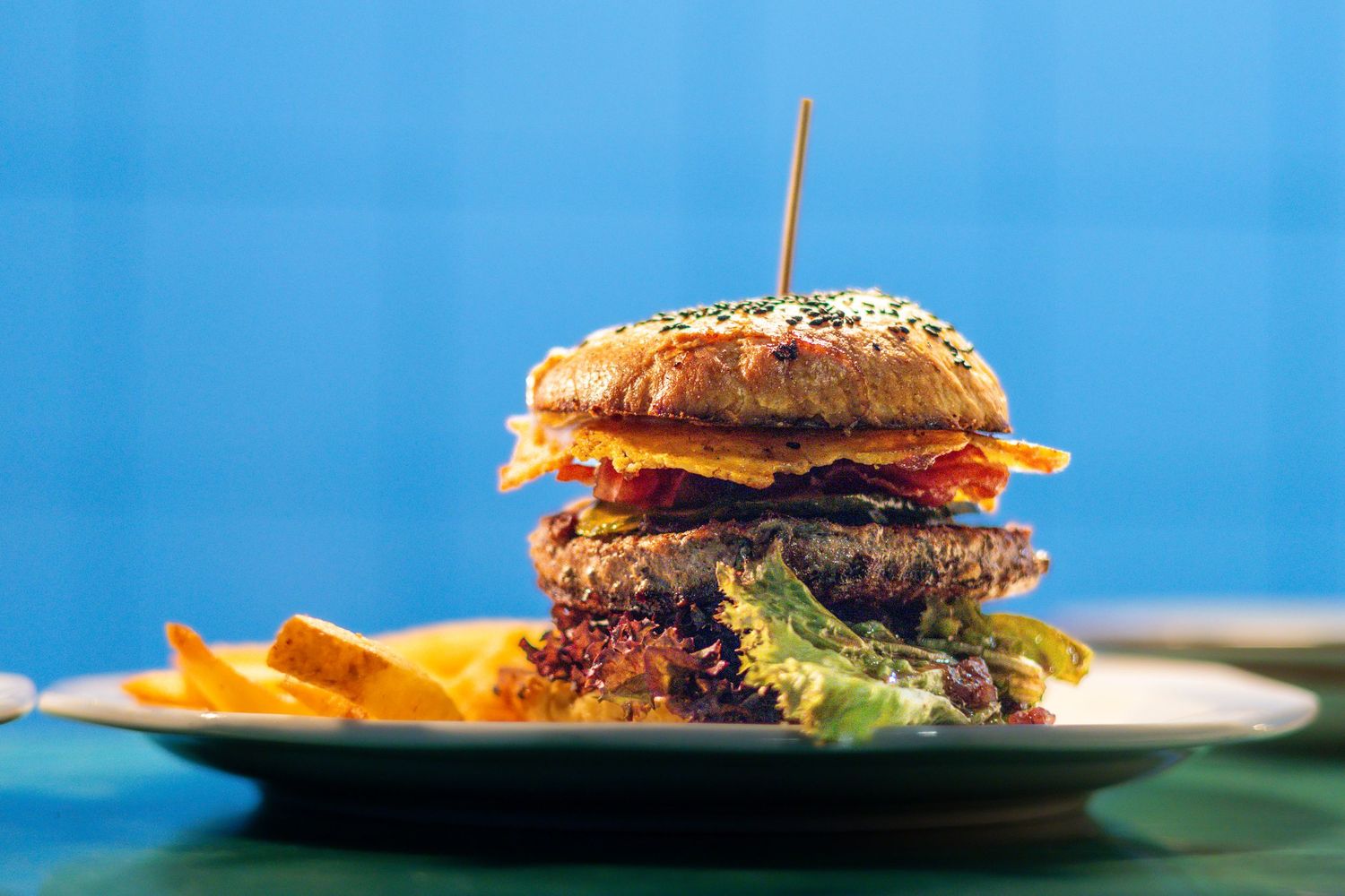 A tall gourmet burger with sesame bun, fresh toppings, and a toothpick, served with fries on a plate.
