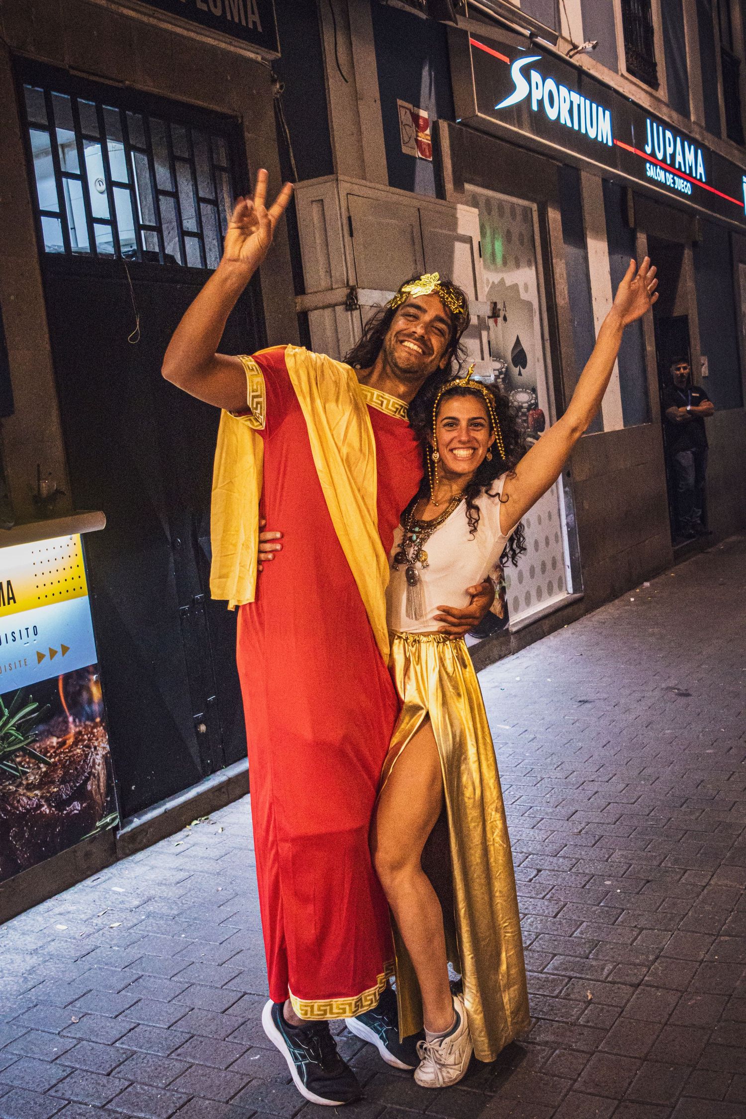 Two people in red and yellow costumes pose playfully on a city street at night outside storefronts.