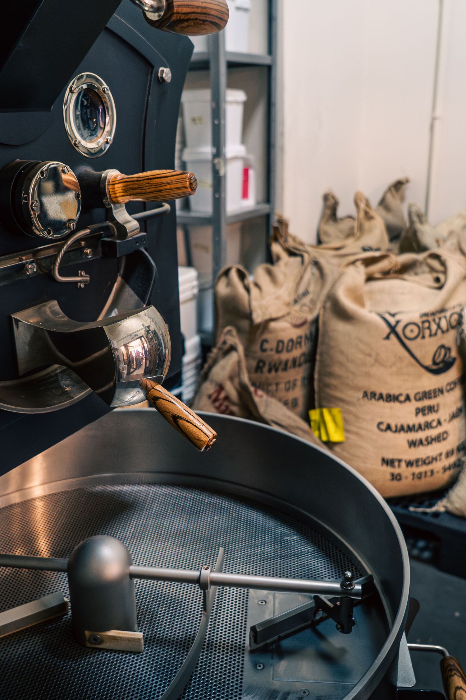 Coffee roaster machine with cooling tray and burlap sacks of Arabica green coffee beans from Peru in background.