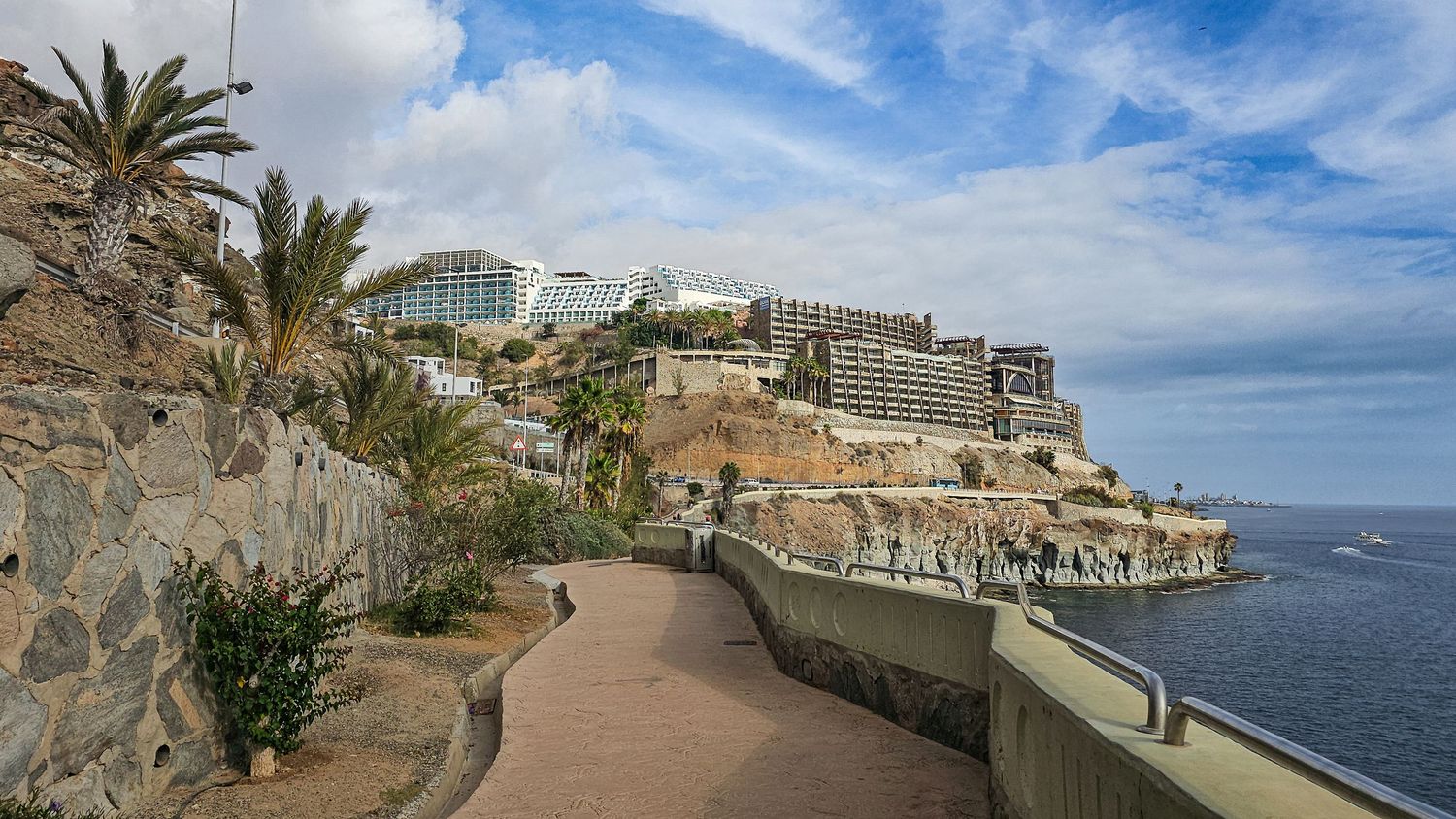 Panoramic view of a coastal walkway with palm trees and cliffside buildings along the Mediterranean Sea under blue skies.