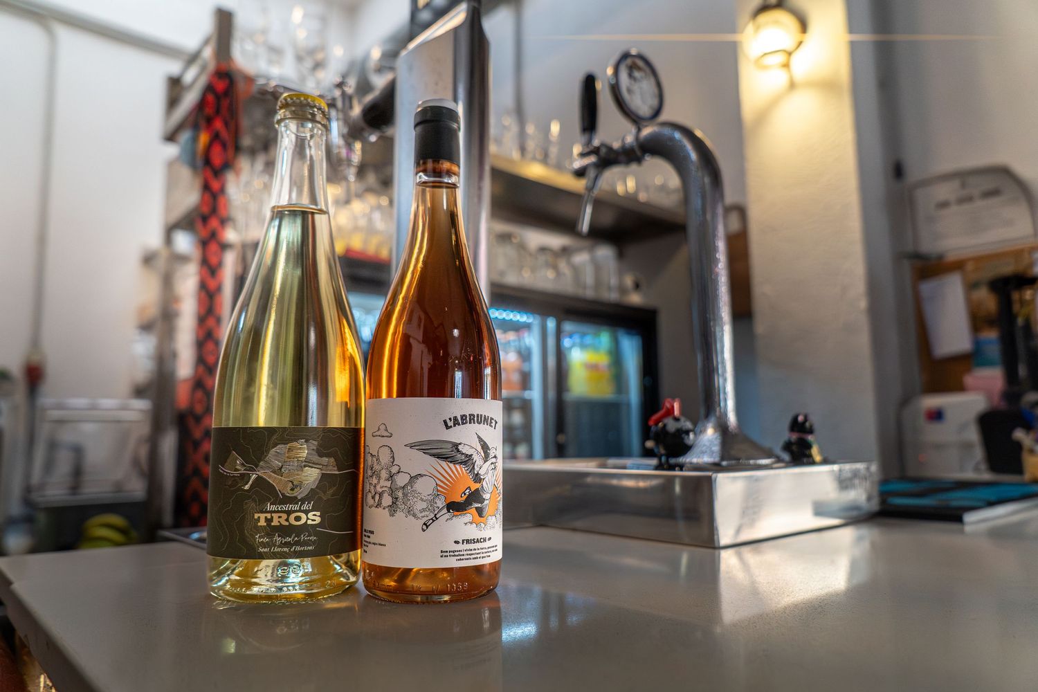 Two wine bottles on a bar counter, one white and one rosé, with tap handles and refrigerator in background.