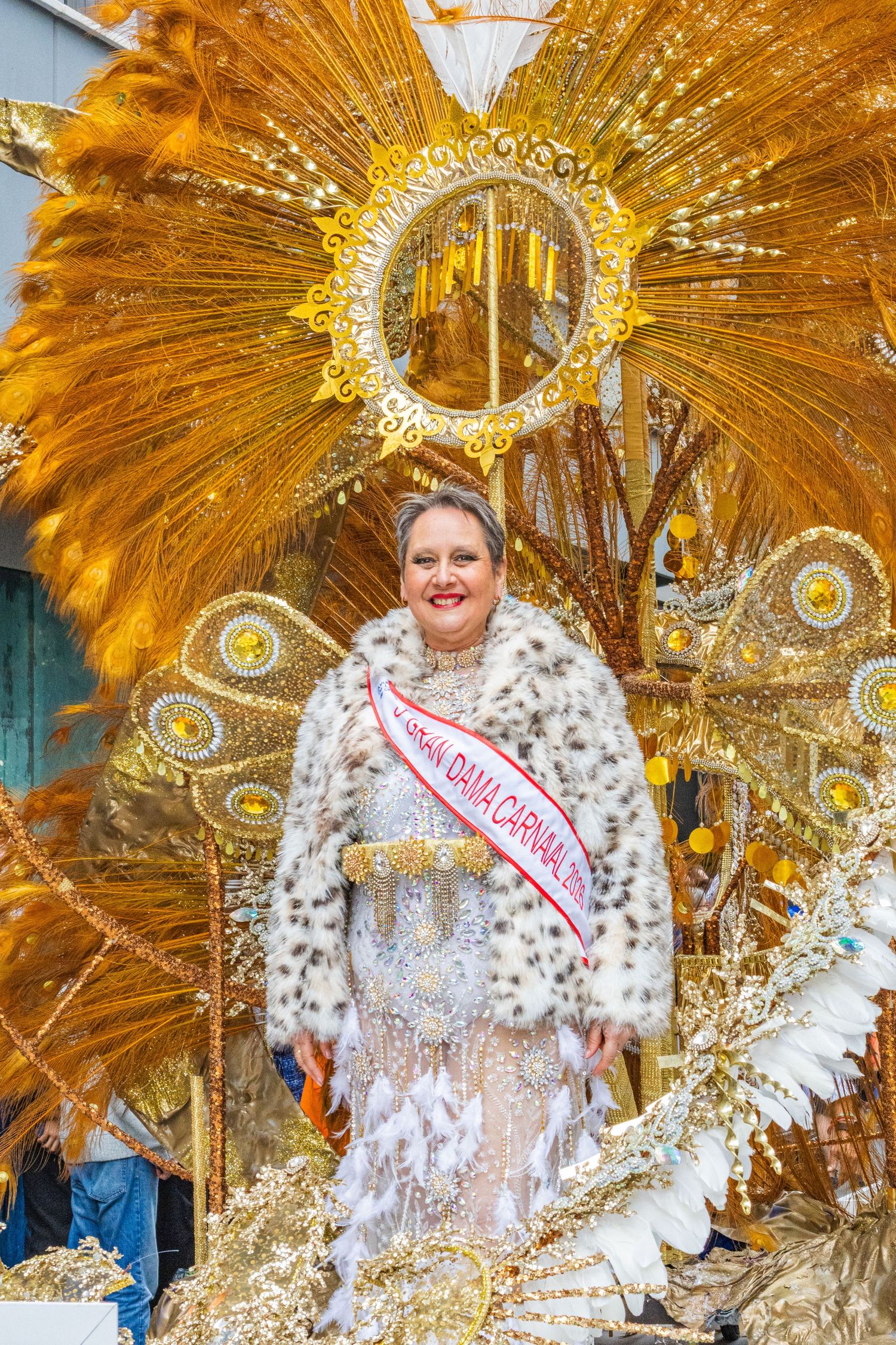 Carnival performer in ornate gold feathered headdress and spotted white costume poses with pageant sash.