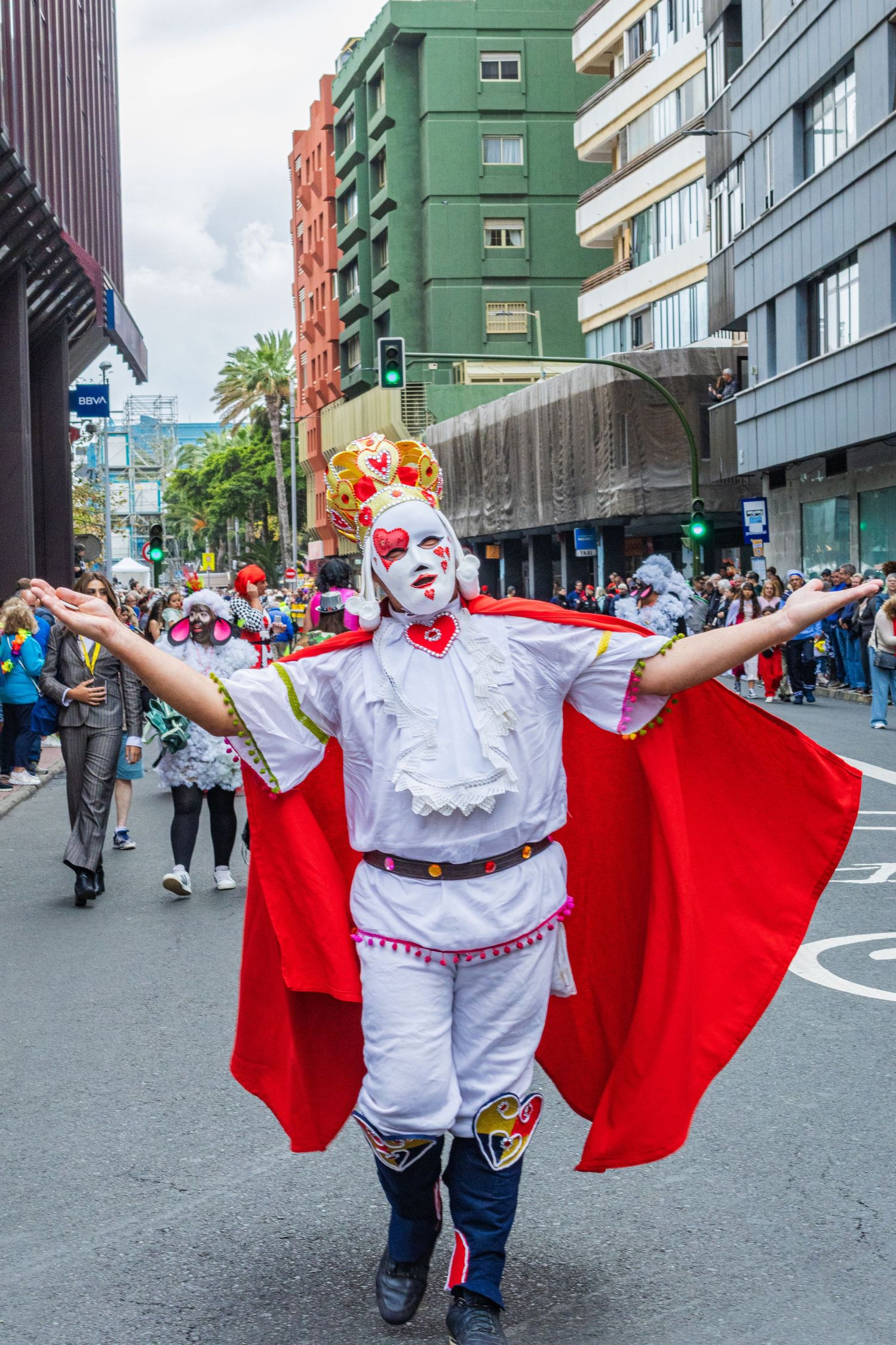 Costumed performer in white and red attire with a cape dances during a street festival in an urban setting.