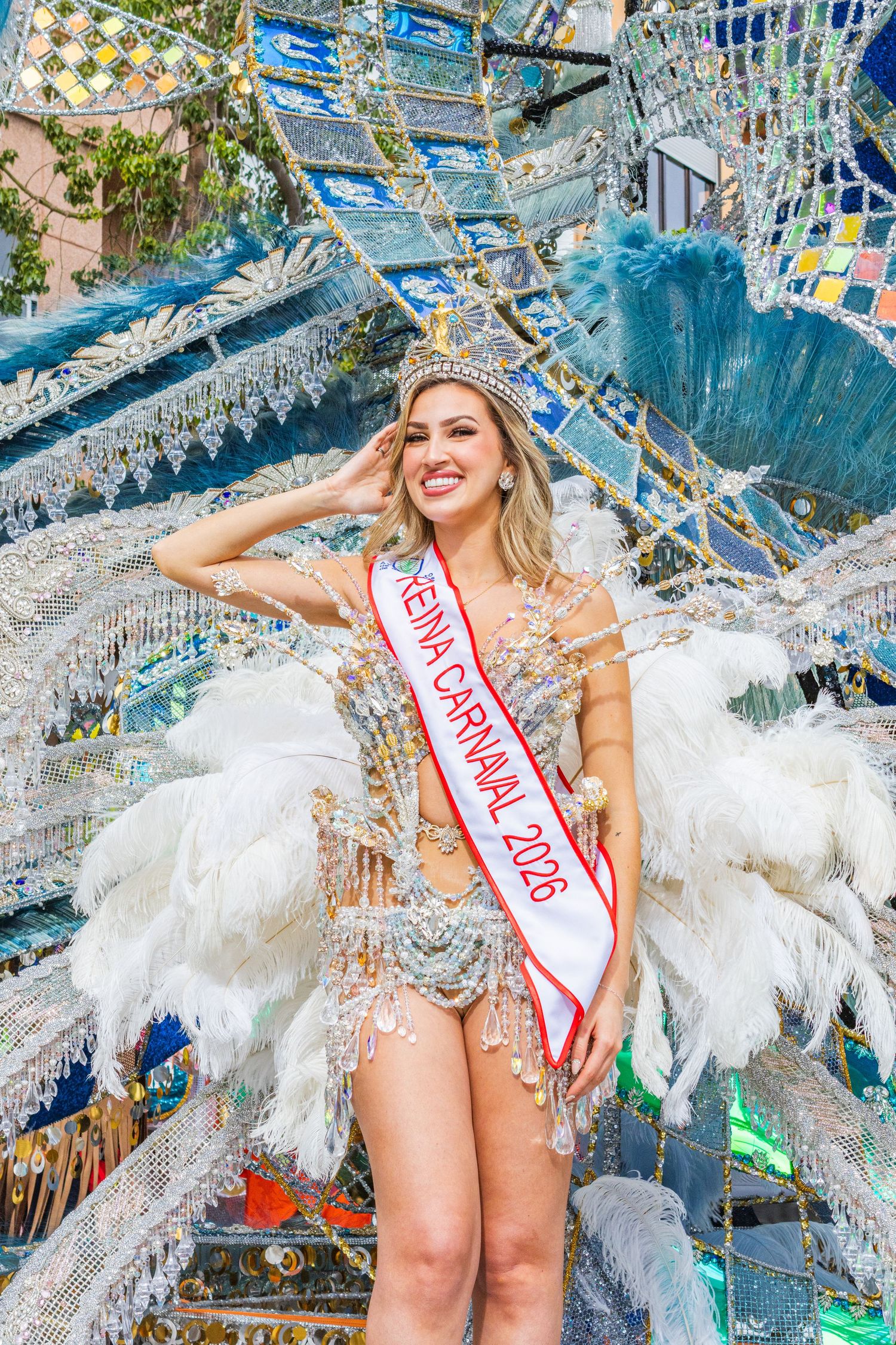 Carnival performer in white bodysuit with feathered wings poses against colorful mosaic wall background.
