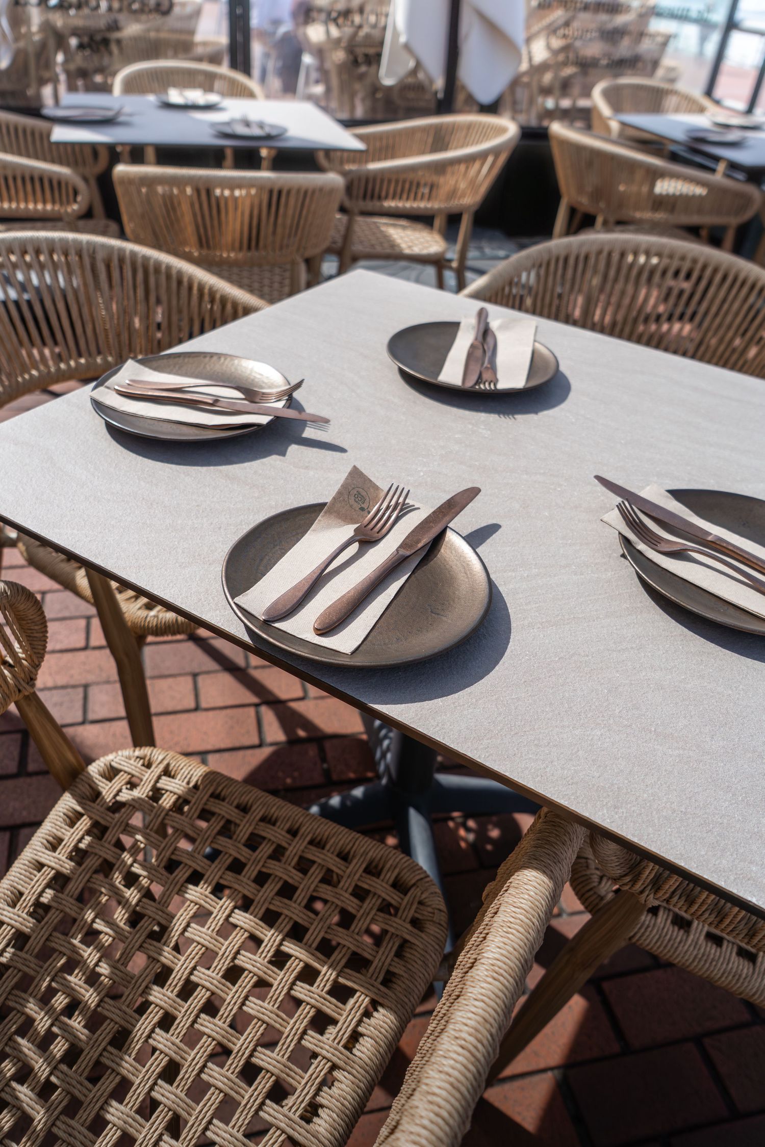 Outdoor restaurant patio with wicker chairs, gray tables set with dark plates, folded napkins, and silverware on brick flooring.