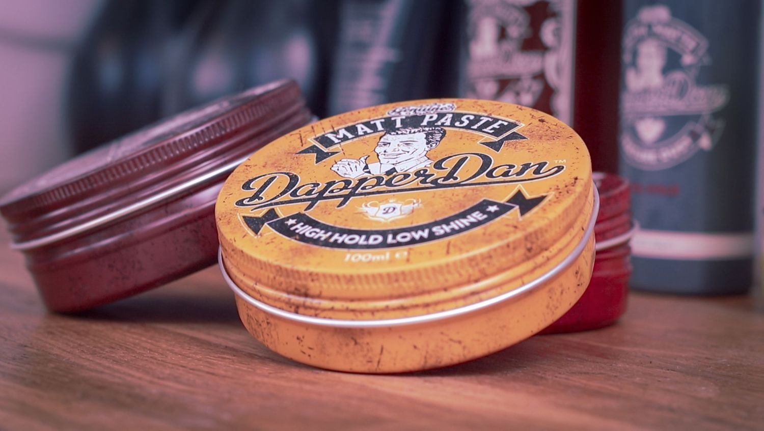A collection of pomade tins and hair styling products arranged on a wooden surface in warm lighting.