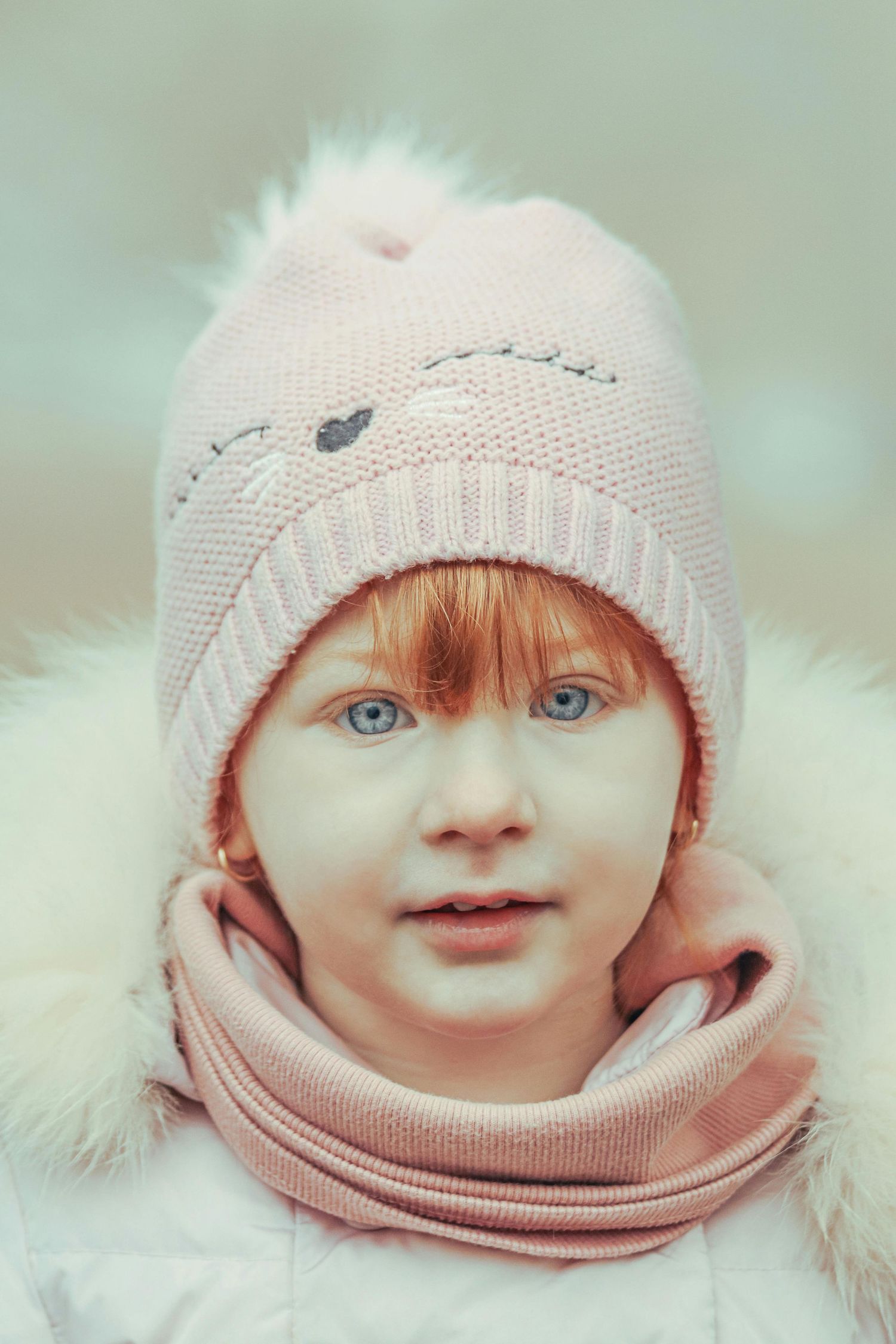Close up portrait of a child wearing a soft pink knit winter hat and scarf against a light background.
