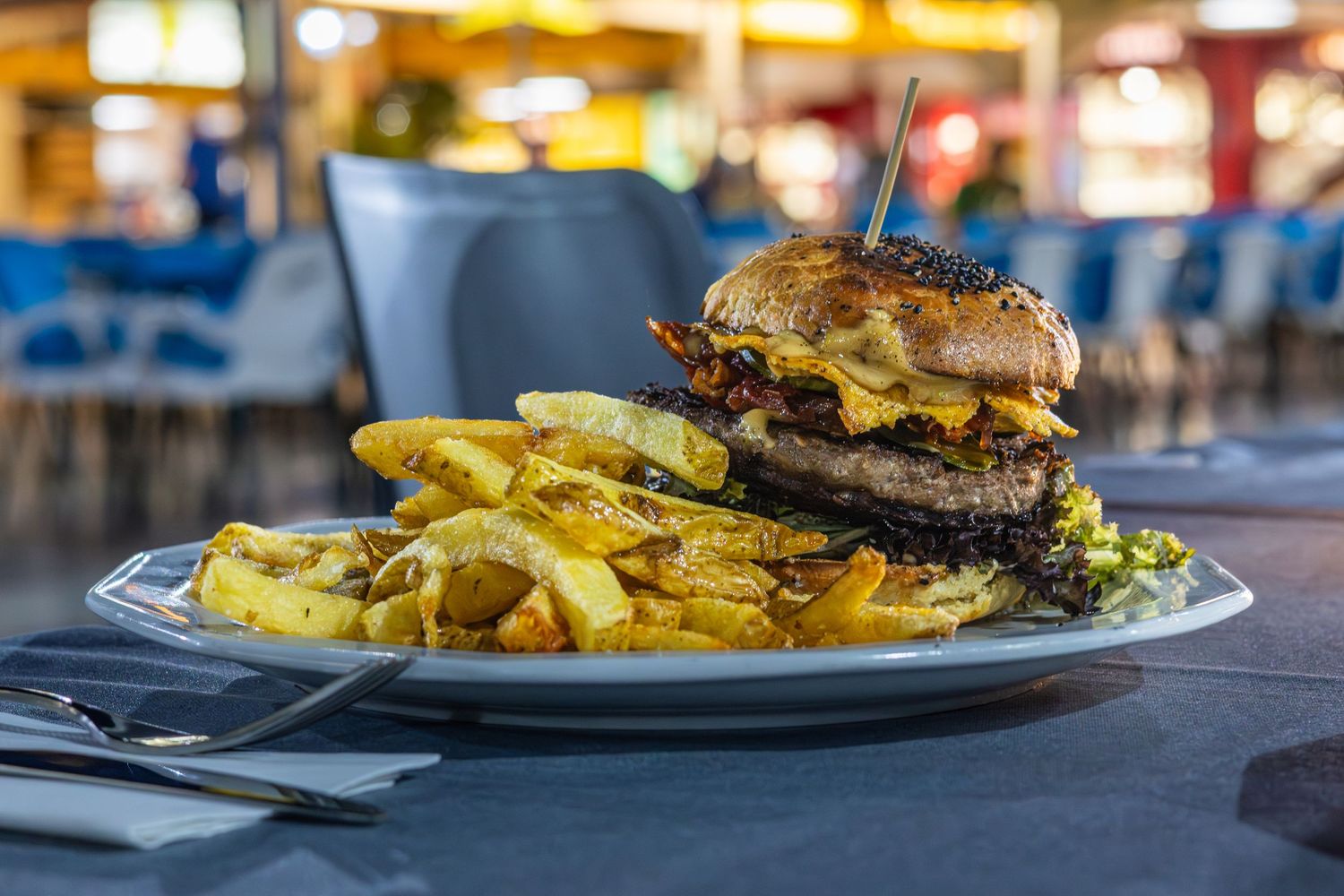 A large double cheeseburger with fries on a white plate at an outdoor restaurant.