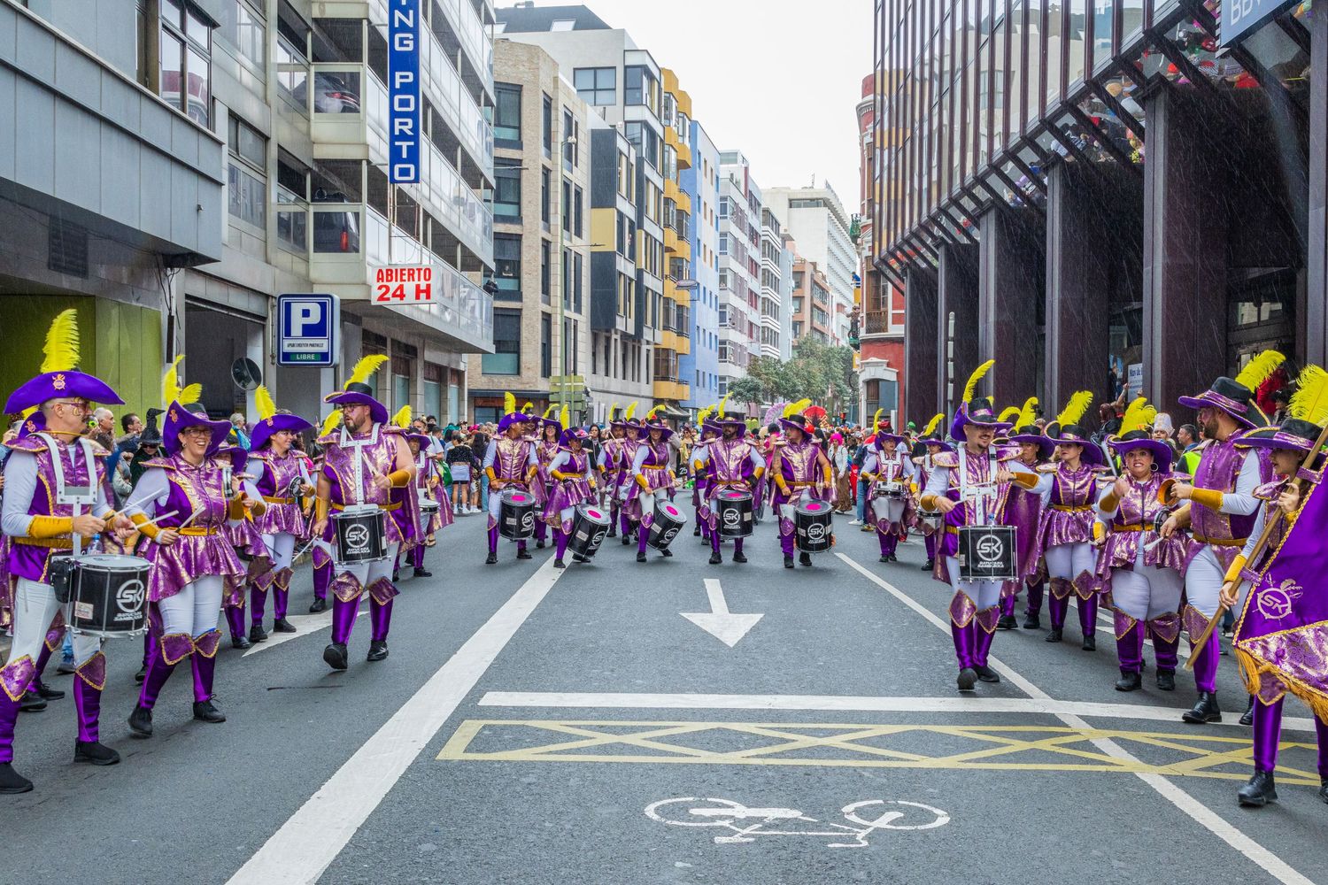 Aerial view of a street parade with performers in purple uniforms marching in formation through a city street.