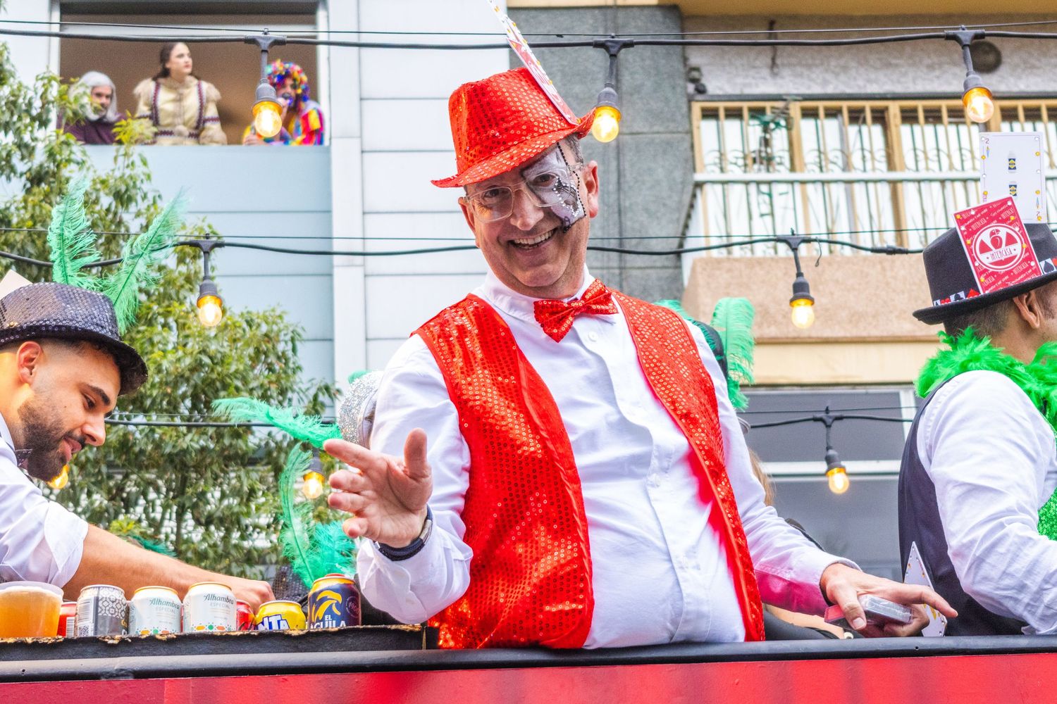 Parade participant wearing bright red vest and bow tie waves from float during festive celebration.