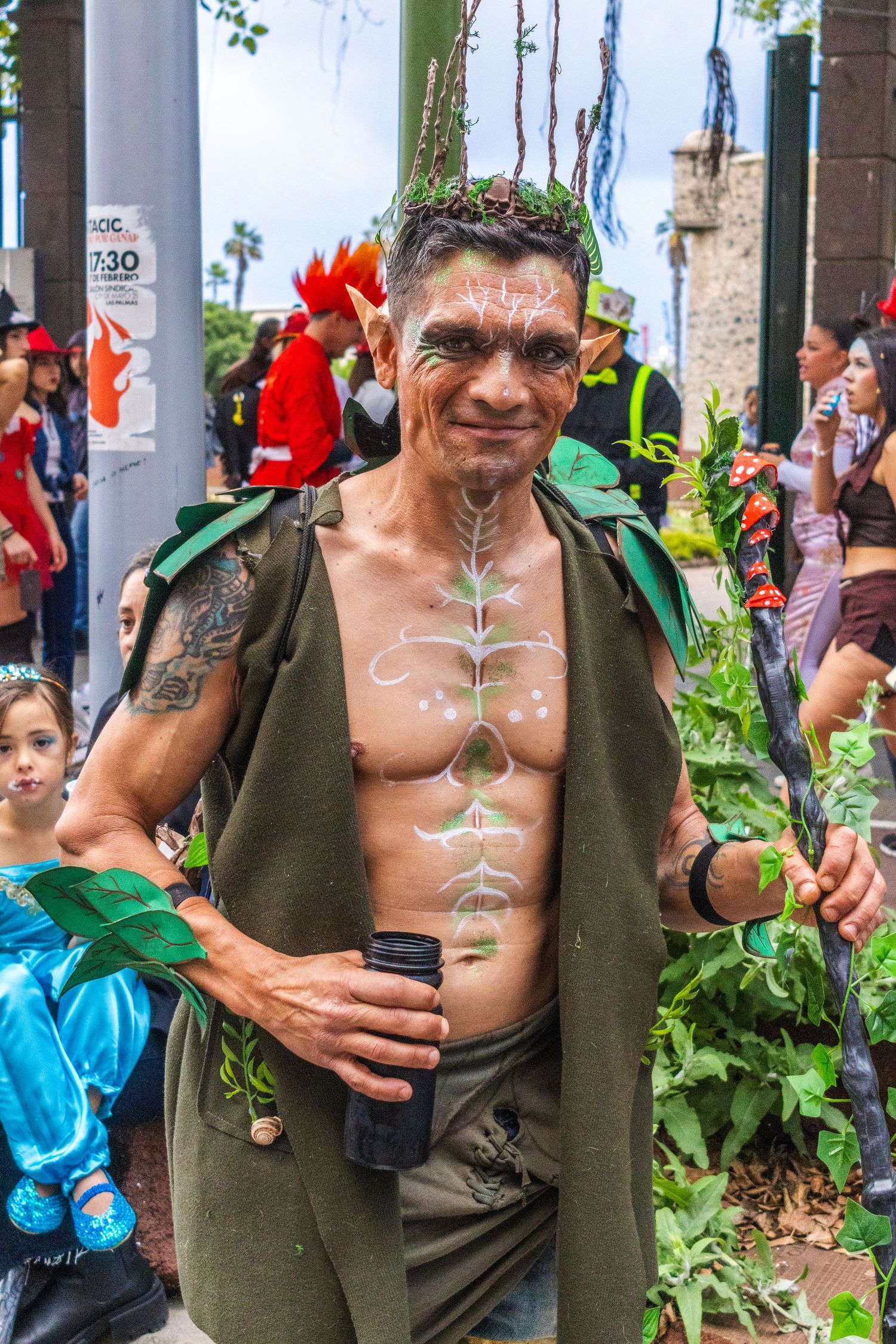 A performer in green costume paint poses with plants and flowers at an outdoor festival.