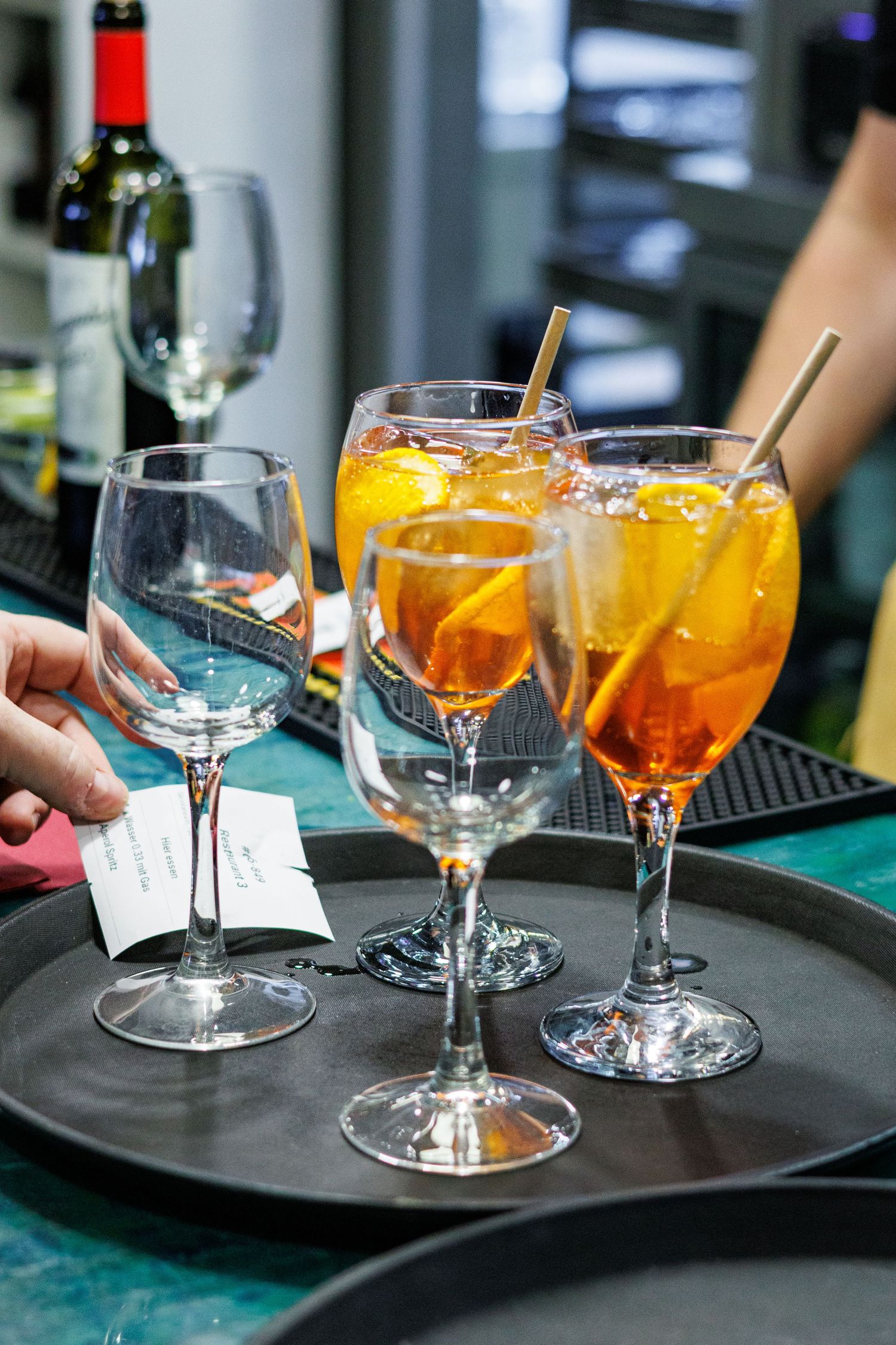 Two Aperol Spritz cocktails with orange slices and straws on a bar tray beside empty wine glasses.