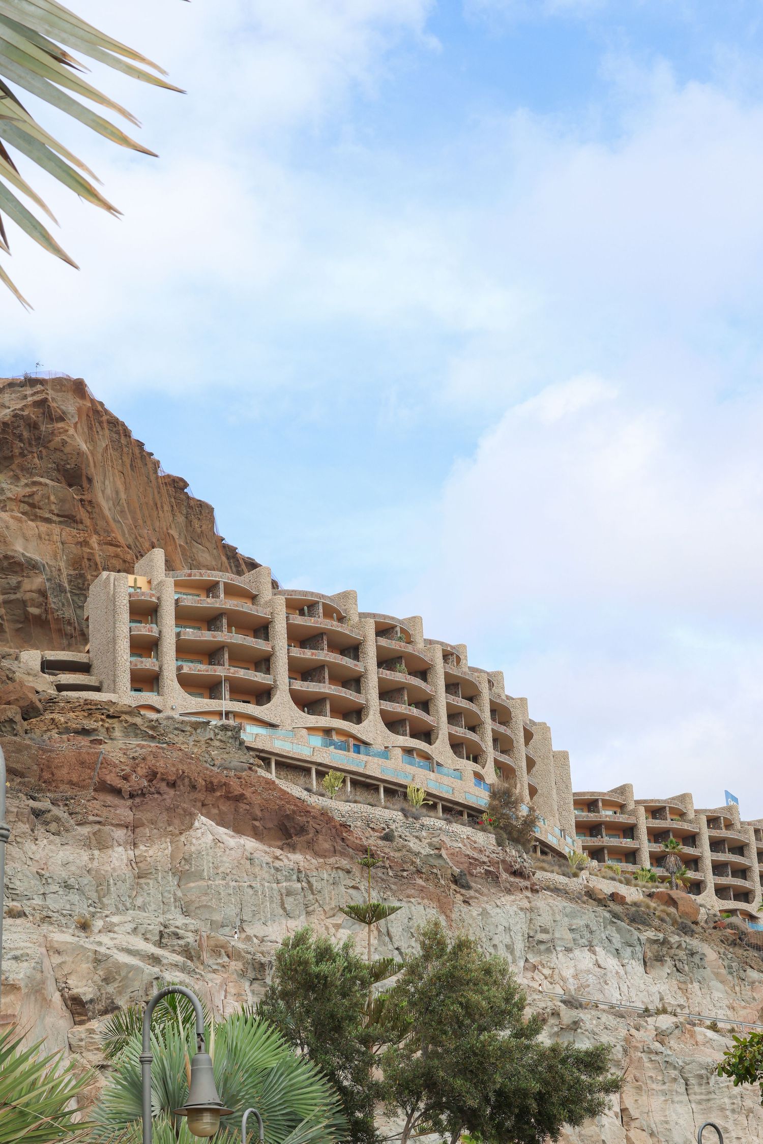 Modern apartment buildings perched dramatically on top of rugged desert cliffs with palm trees below against a blue sky.