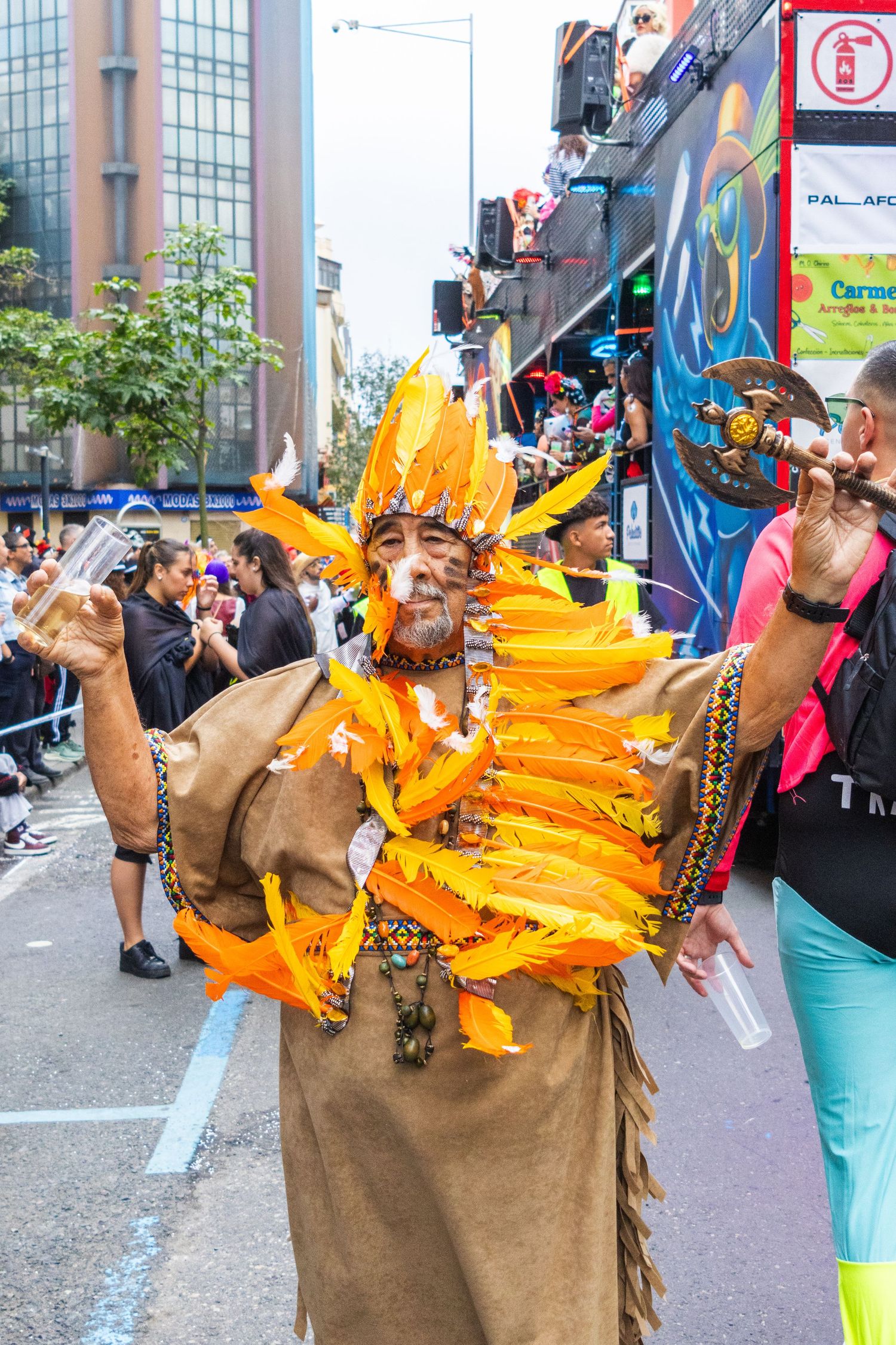 A performer in a brown costume with vibrant orange feathers dances energetically during a street festival celebration.