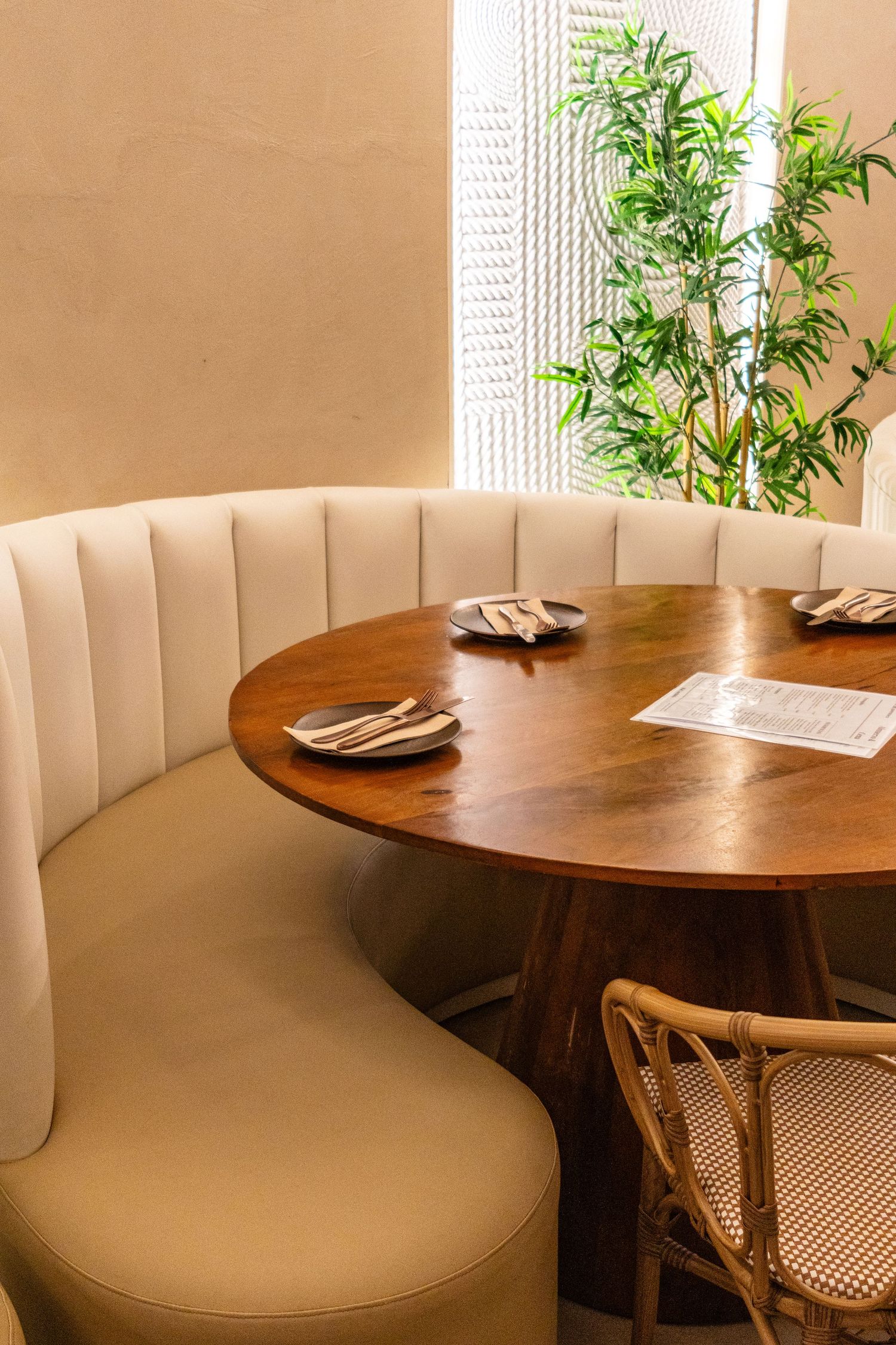 Elegant restaurant booth with cream tufted banquette seating, round wood table, place settings, and a green plant by the window.