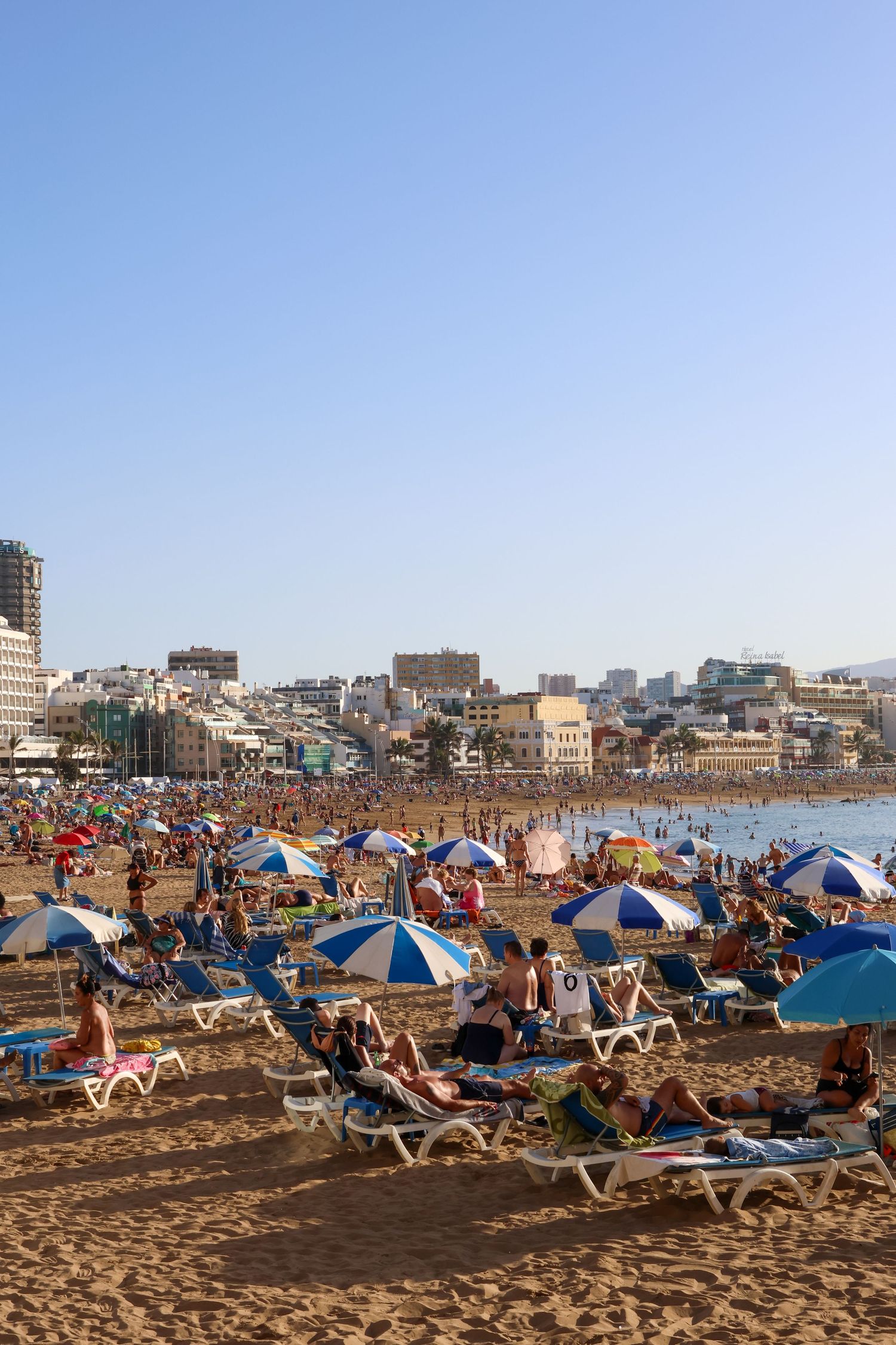 Crowded beach scene with blue and white umbrellas, lounge chairs, and city buildings visible in the background.