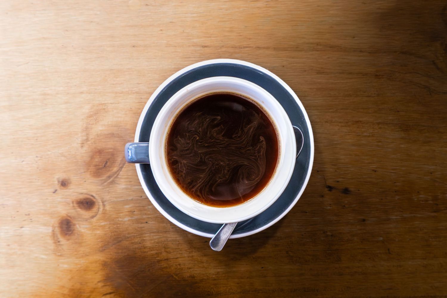 Overhead view of a black coffee in a striped cup and saucer with a spoon on a wooden table.