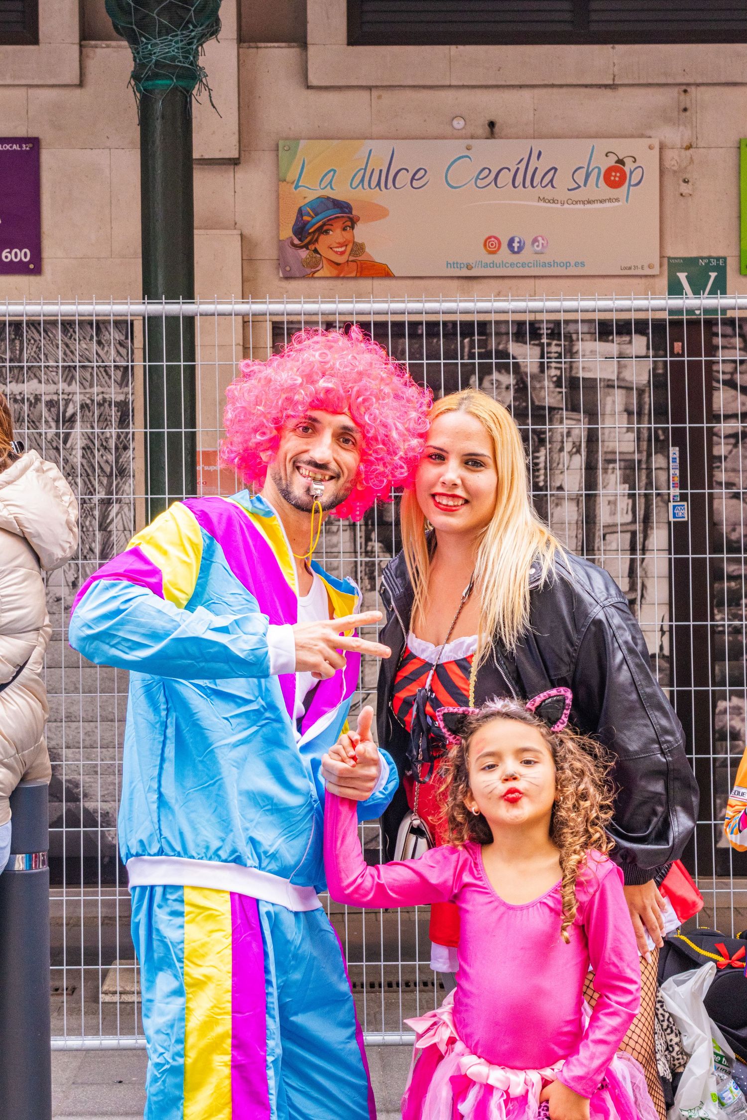 Performers in bright blue and pink costumes with colorful wigs pose playfully on a city street.