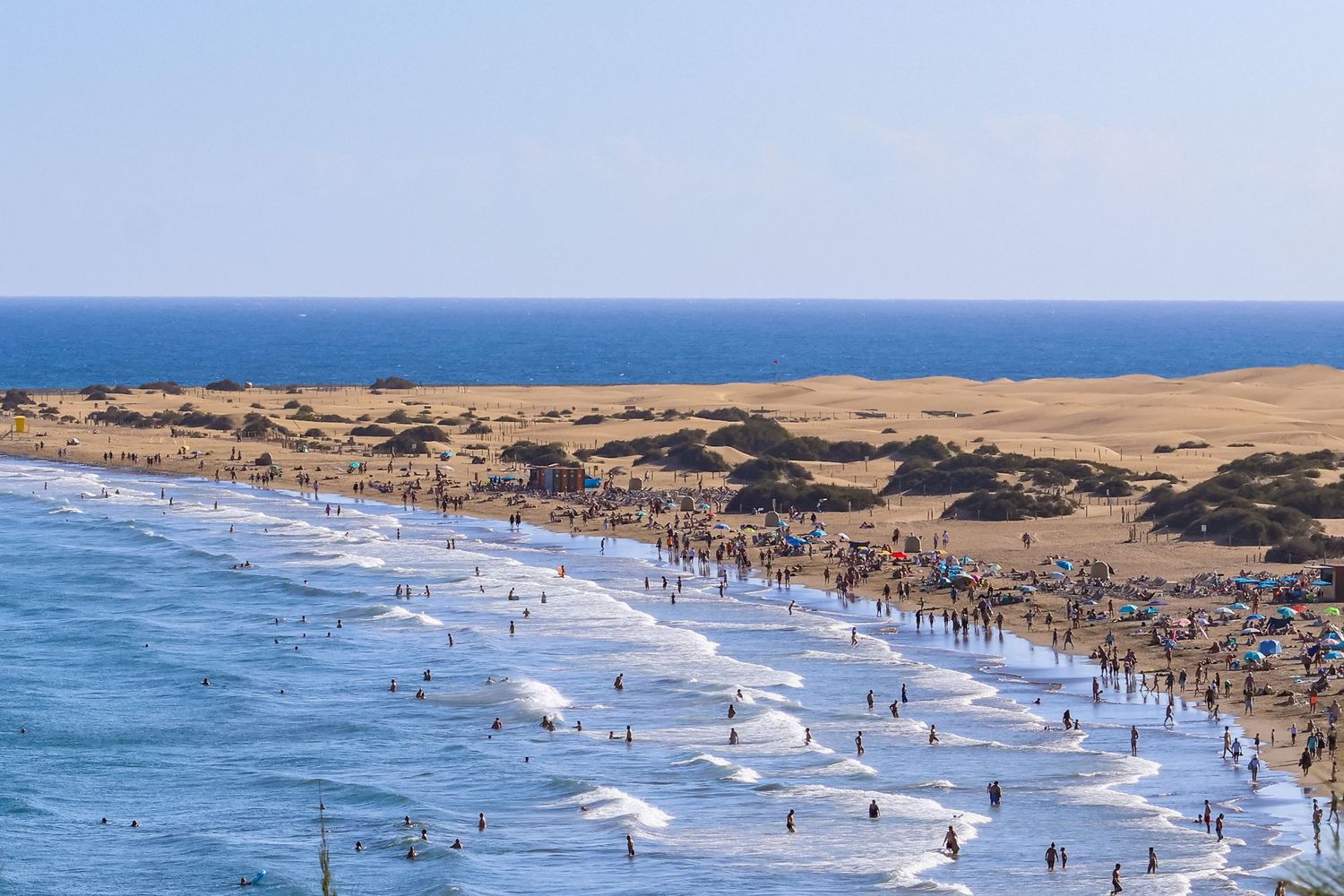Aerial view of crowded beach with surfers riding waves near sand dunes and blue ocean in summer.