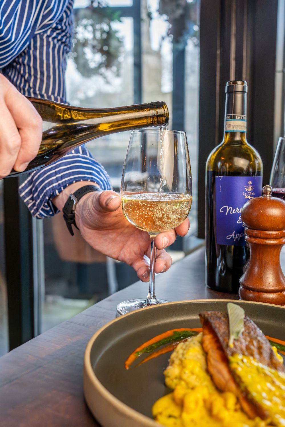 White wine being poured into a glass next to a plated dish with yellow risotto and meat on a restaurant table.