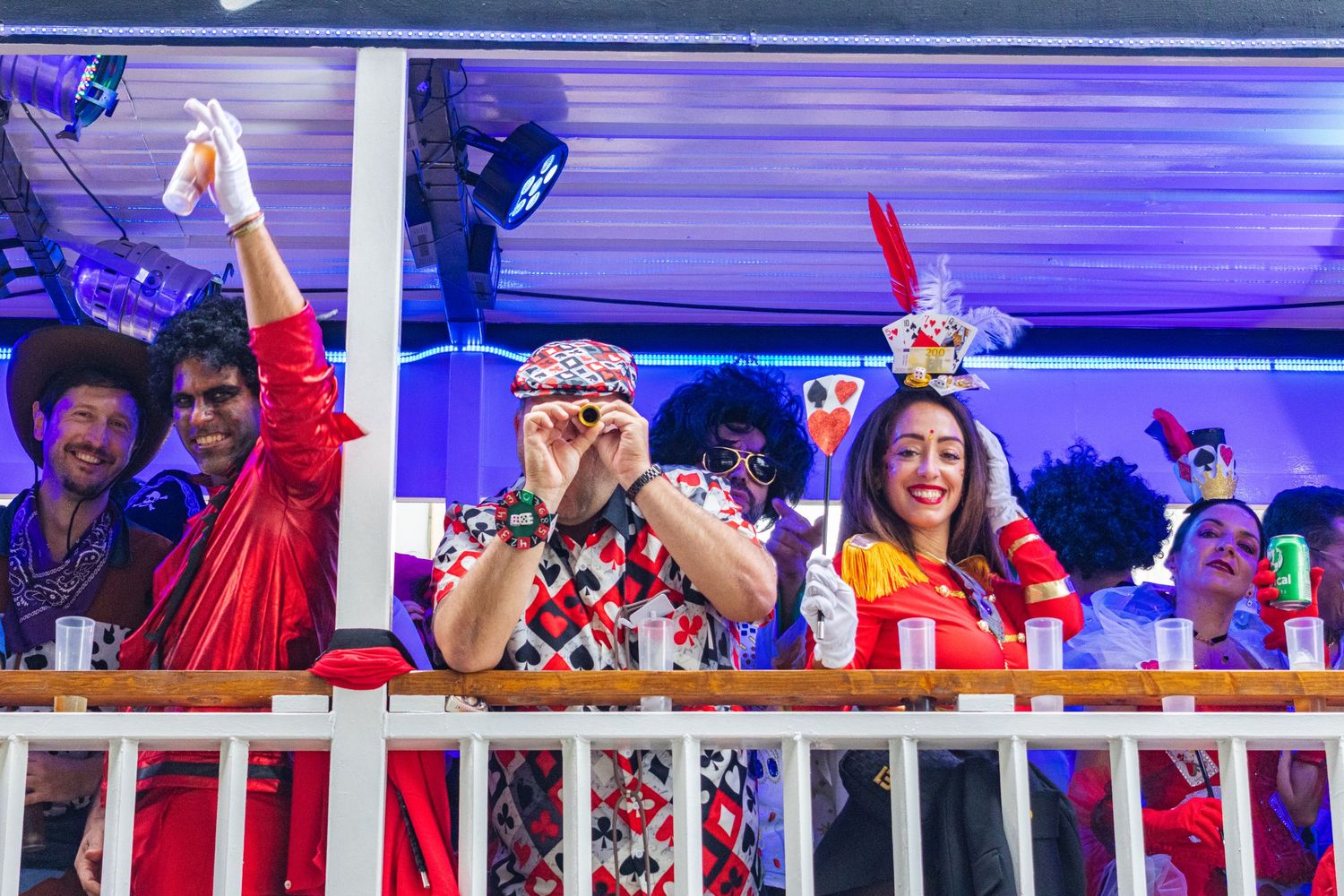 Festive partygoers in colorful outfits and bunny ears celebrate from a balcony at a nighttime event.