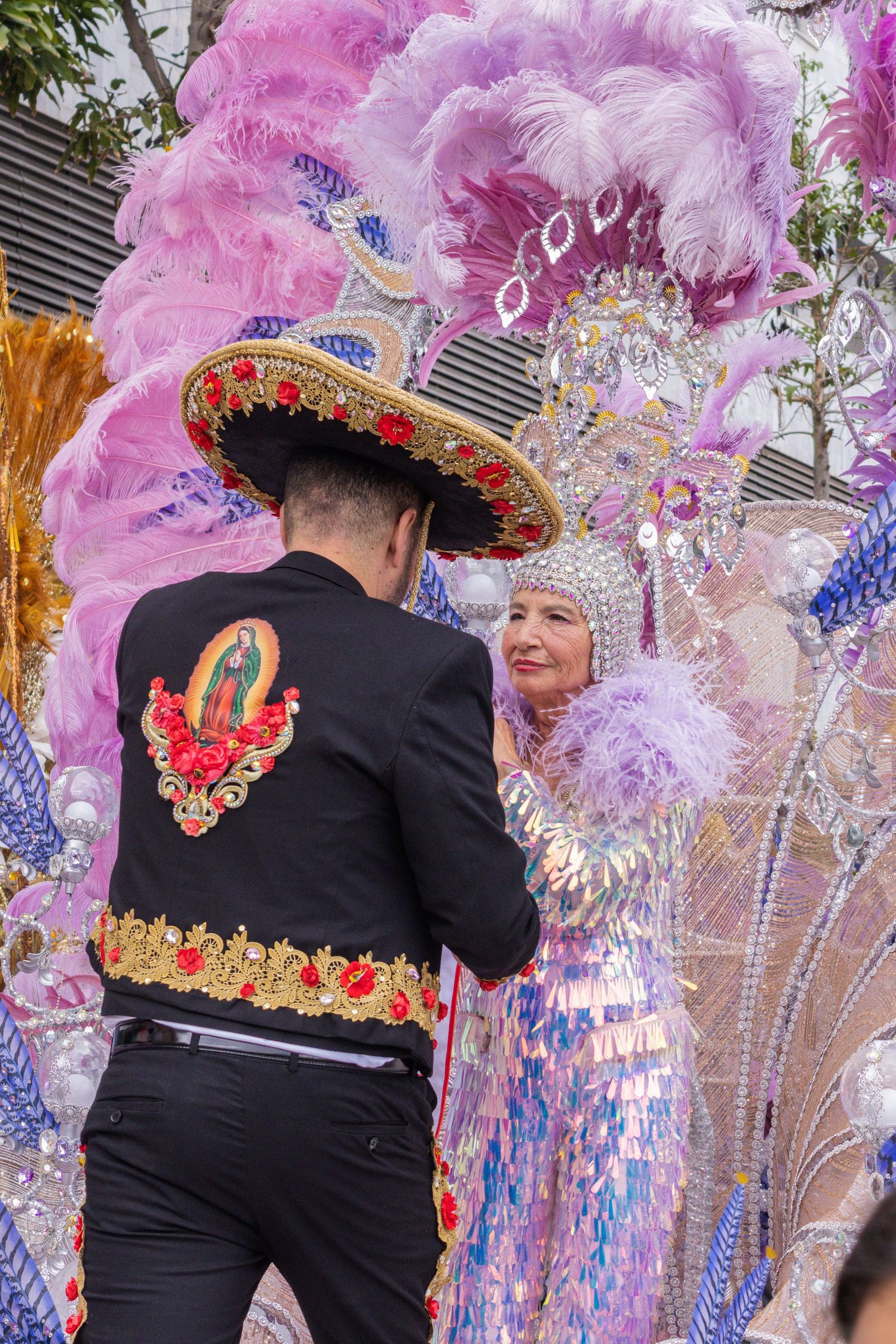 Carnival performers in black costumes with ornate headdresses adorned with pink and purple feathers dance at a festival.