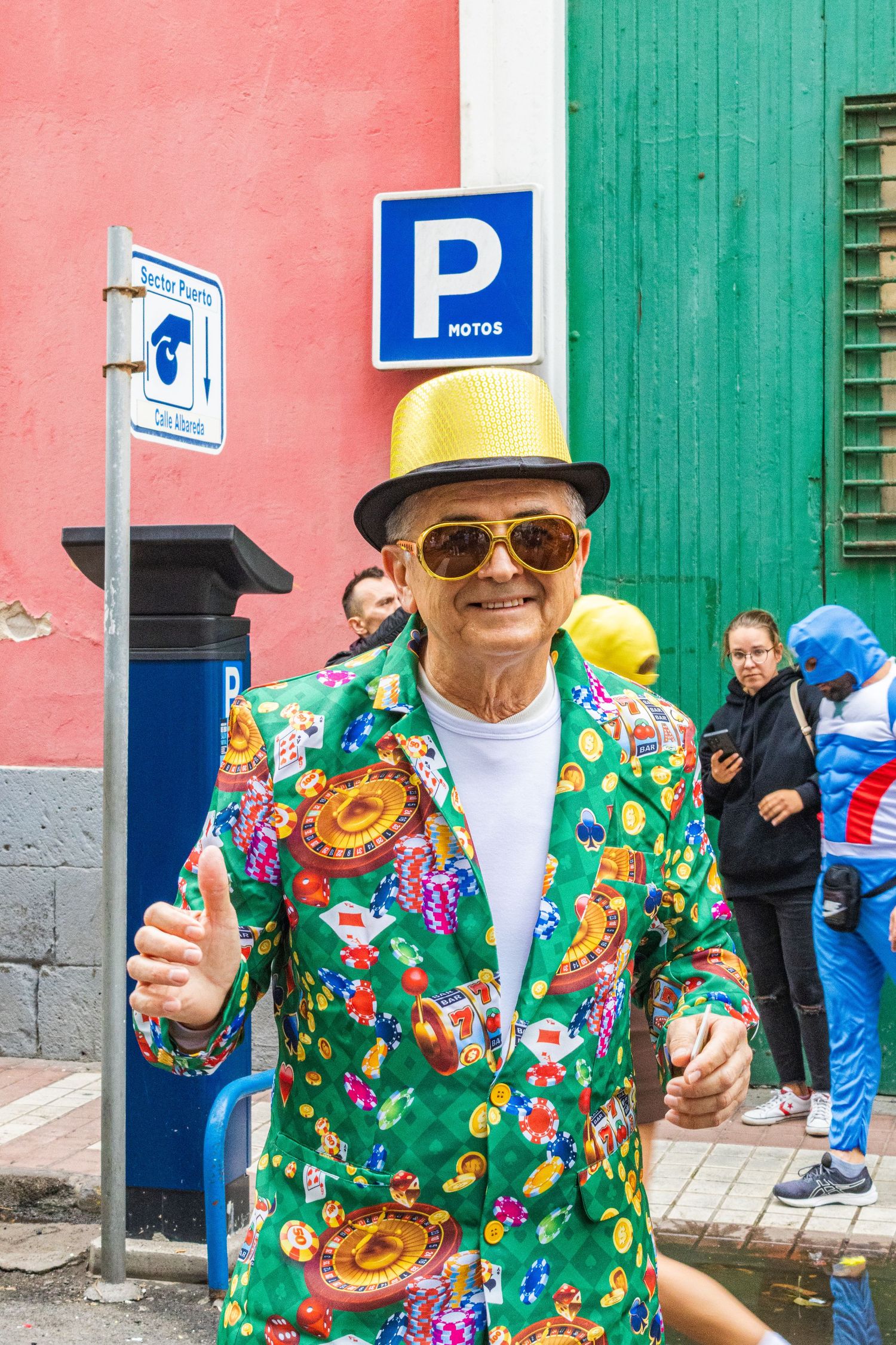 Person wearing colorful floral Hawaiian shirt and sunglasses poses with a drink against pink and green wall.