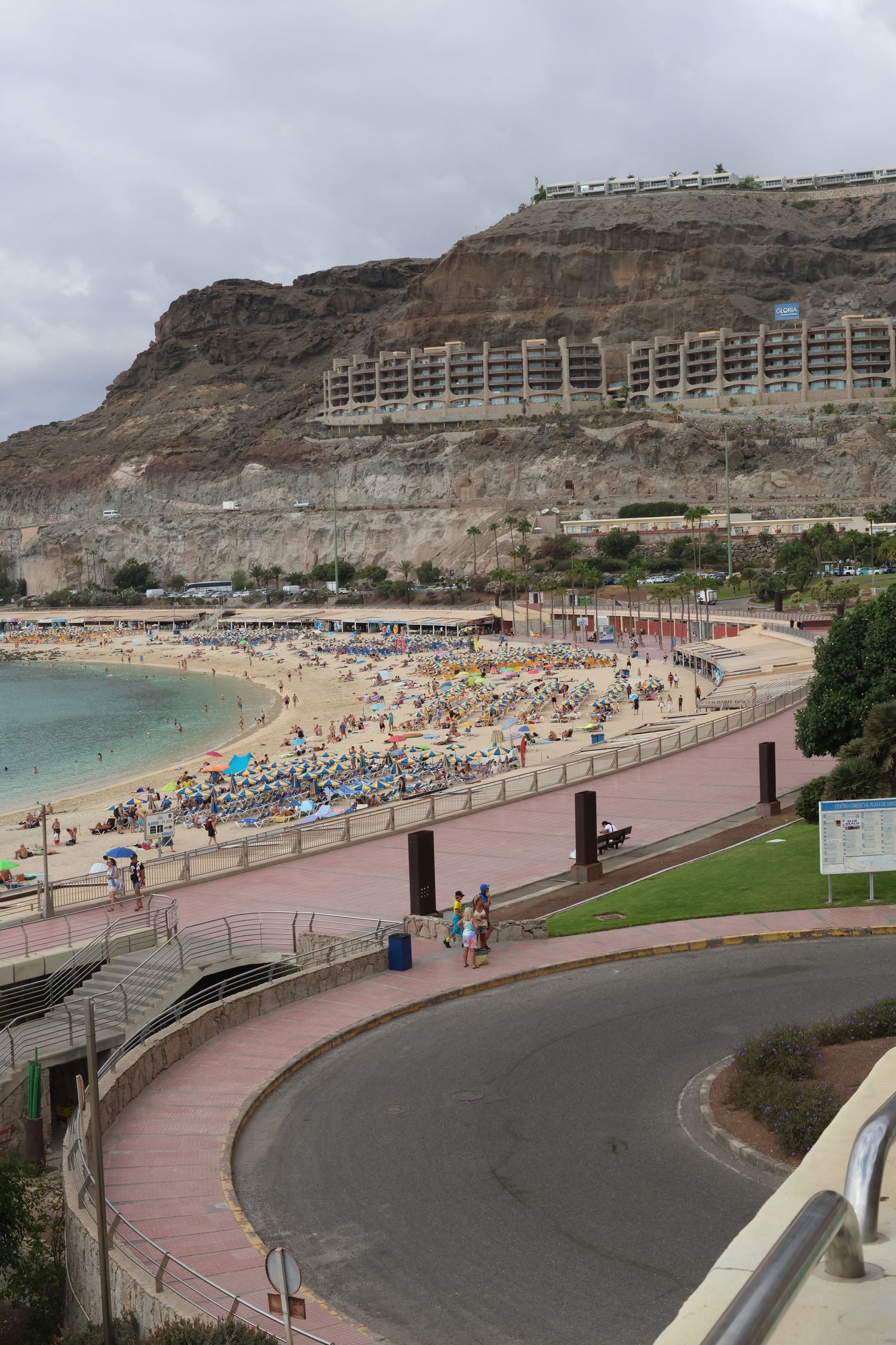 Scenic view of a crowded beach with cliffside buildings and a curved promenade walkway in Gran Canaria.
