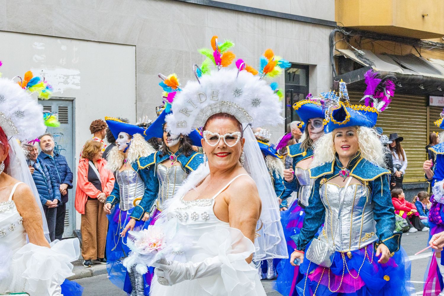 Colorful parade performers in white and blue costumes with feathered headdresses dance during a festive celebration.