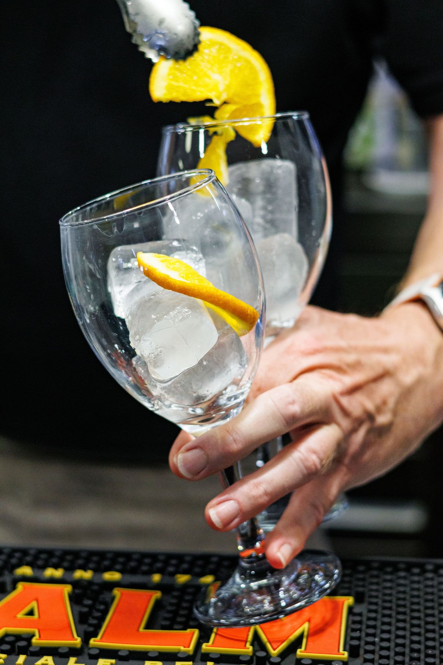 Bartender preparing two elegant balloon gin glasses with ice cubes and fresh orange peel garnish on a bar counter.