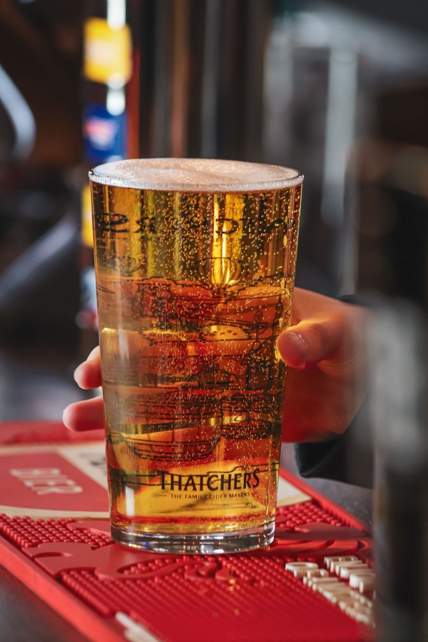 A glass of golden Thatcher's cider sits on a red bar mat, capturing the light and bubbles in the amber liquid.