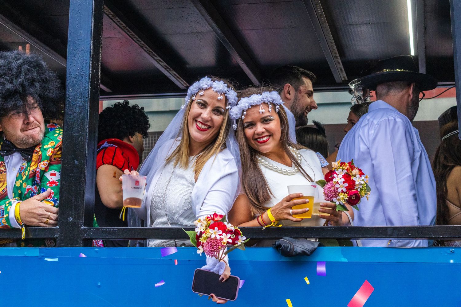 Festival goers in white outfits and flower crowns enjoy drinks and celebrations at an outdoor event.