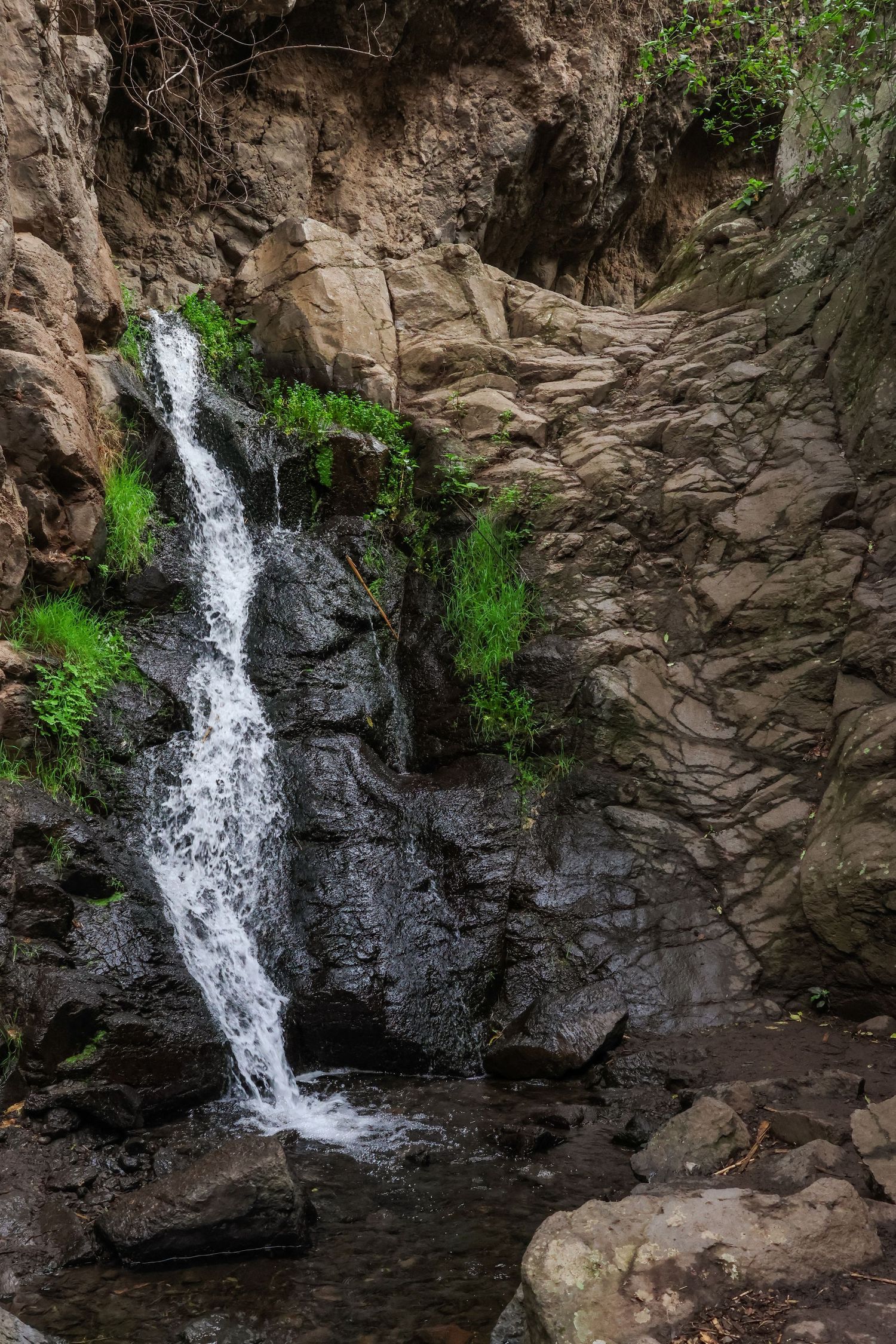 A cascading waterfall flows down rocky cliffs surrounded by green vegetation in a natural canyon setting.