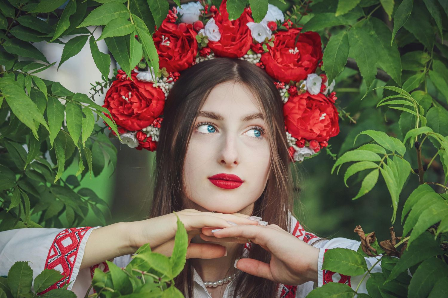 Woman wearing a floral crown of red roses poses against lush green foliage background.